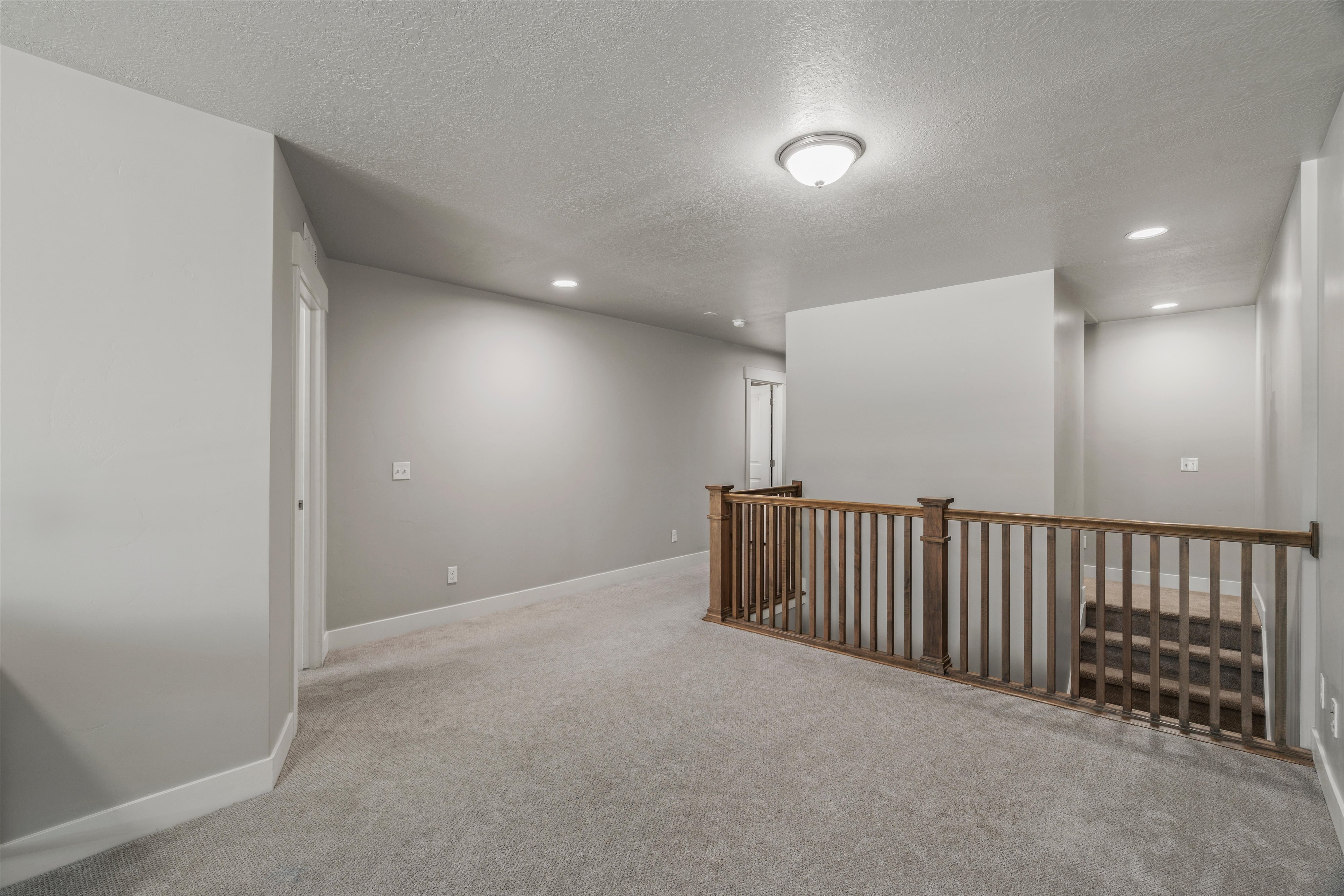 Spacious hallway featuring light gray walls, plush carpet flooring, a natural wood-finish railing with balusters, recessed lighting, and a ceiling-mounted fixture