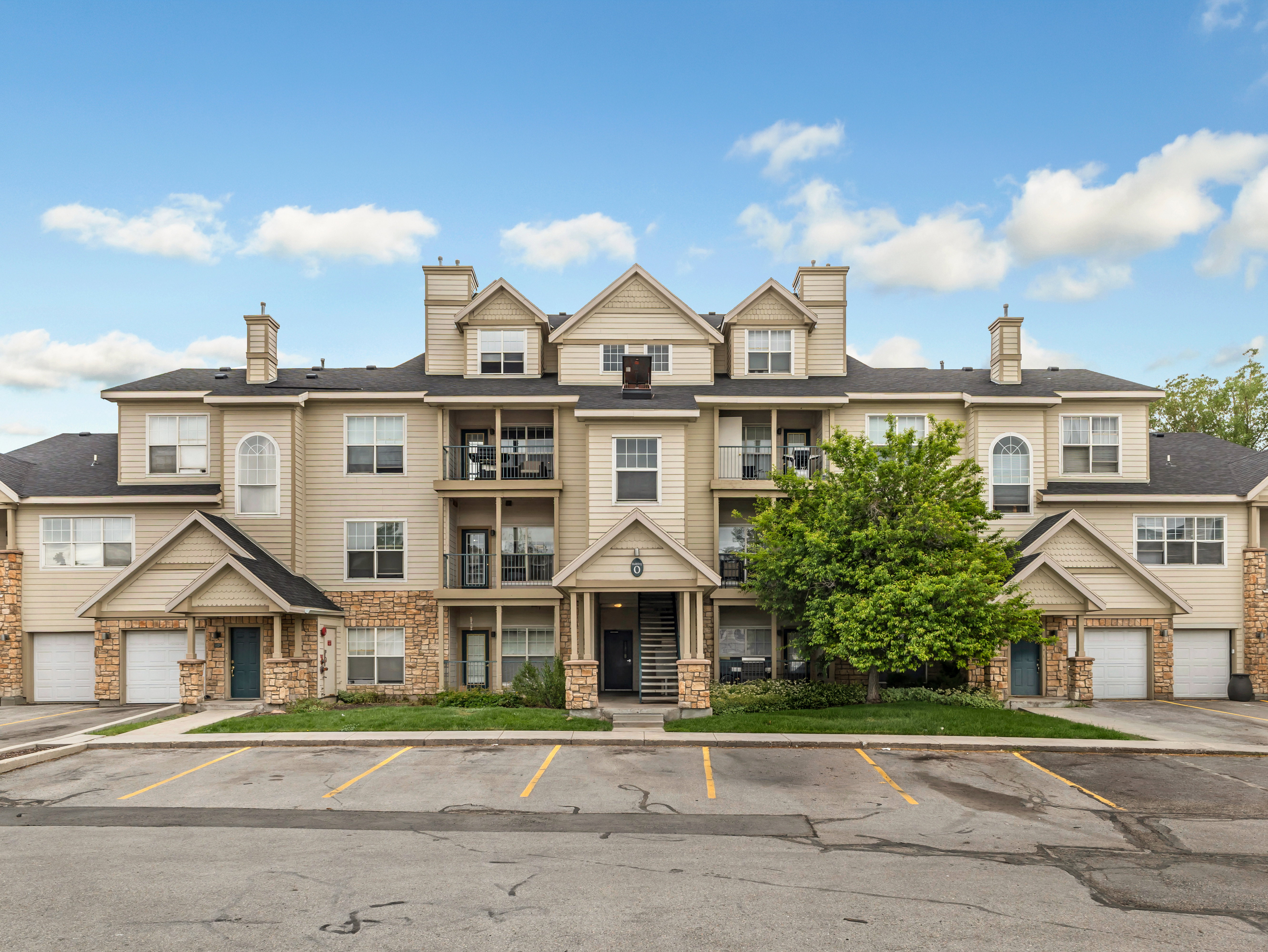 Multi-story residential building featuring light-colored siding, stone accents, multiple balconies with black railings, individual garage doors, and dormer windows