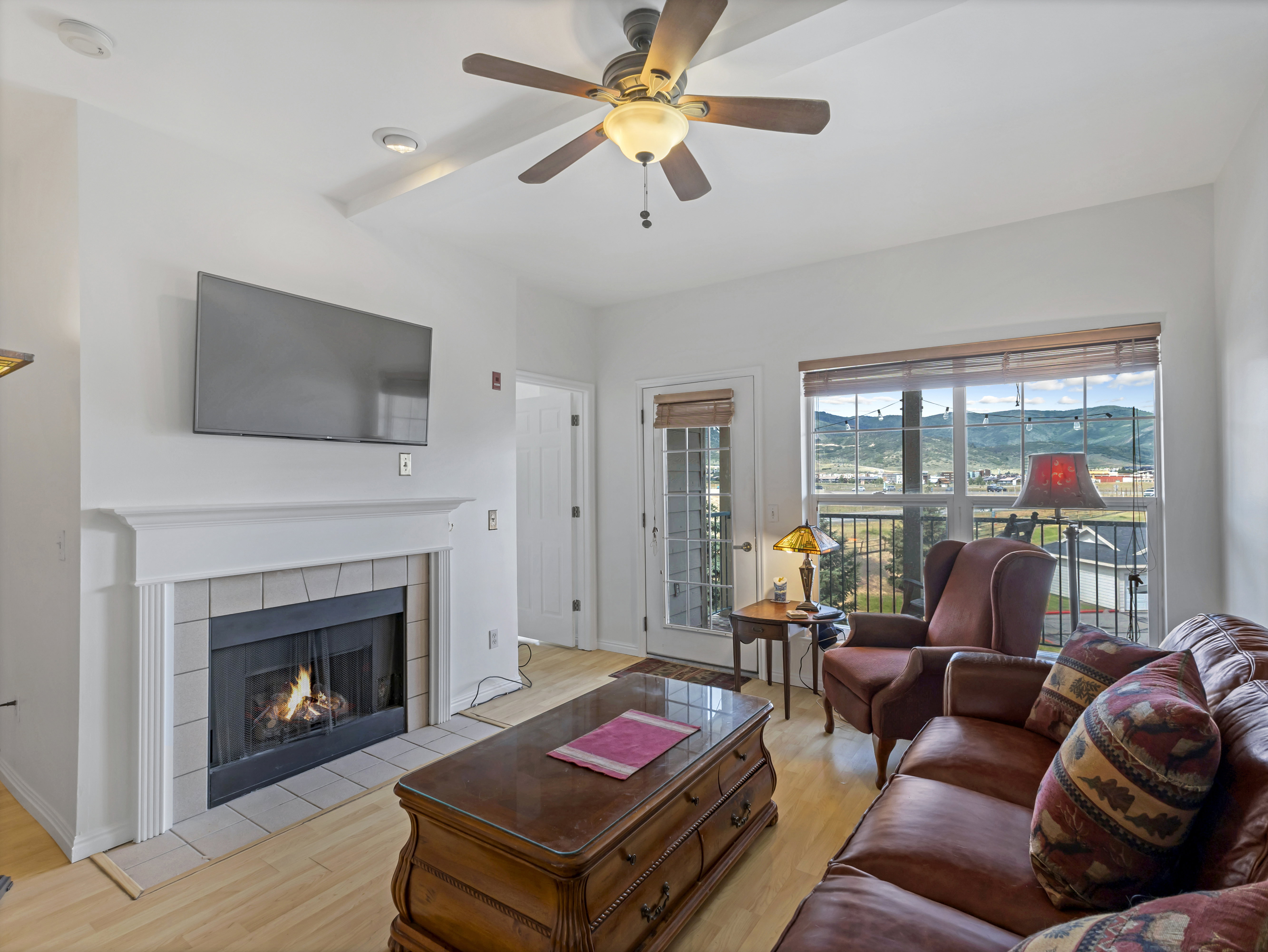 Living area featuring a fireplace with a tiled surround and white mantel, wood-finish flooring, a ceiling fan with integrated lighting, and a glass-paneled door opening to an outdoor space