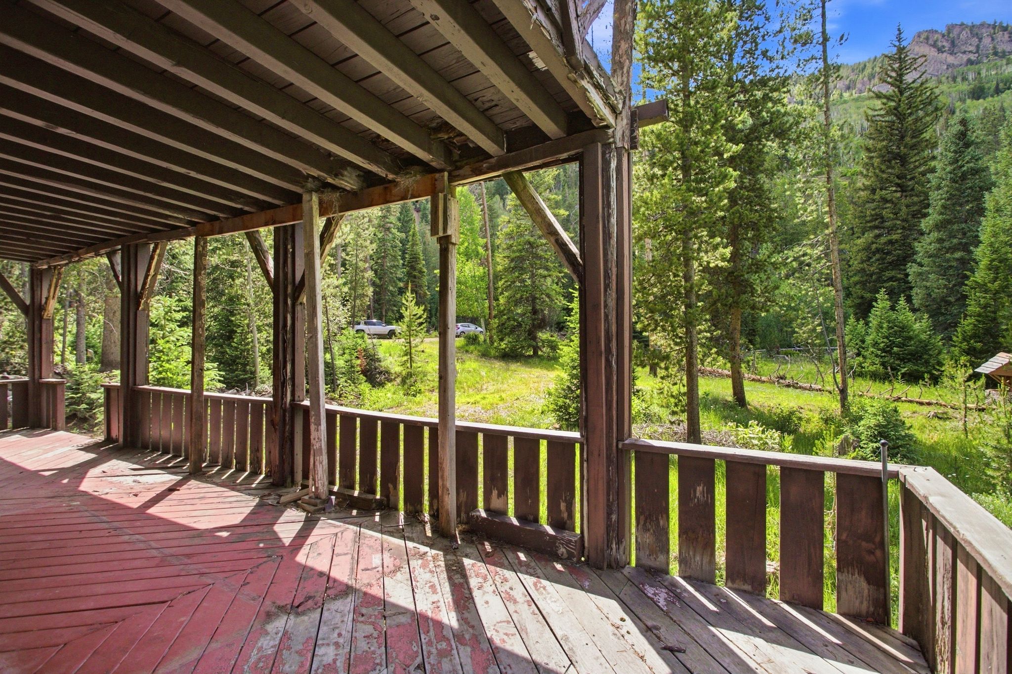 Expansive covered porch with a red-painted wood deck, wooden railing, and structural wood posts, providing an outdoor living space amidst a forested mountain setting