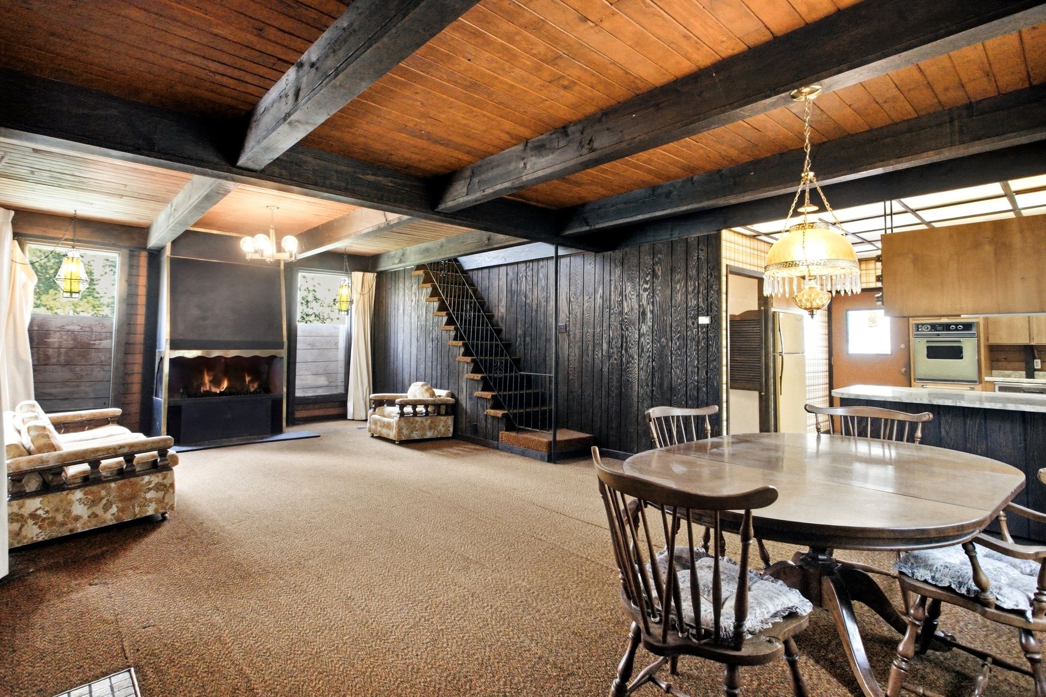 Spacious living area featuring exposed wood beam ceilings, textured dark wood paneling, an integrated fireplace unit, a staircase with open risers, and a dining area with a brass chandelier
