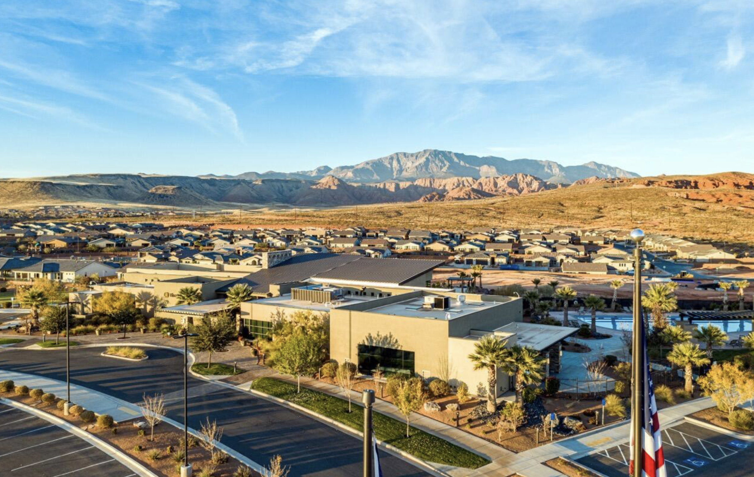 Expansive community featuring desert landscaping, paved roadways, multiple buildings with large windows, and a mountain range backdrop