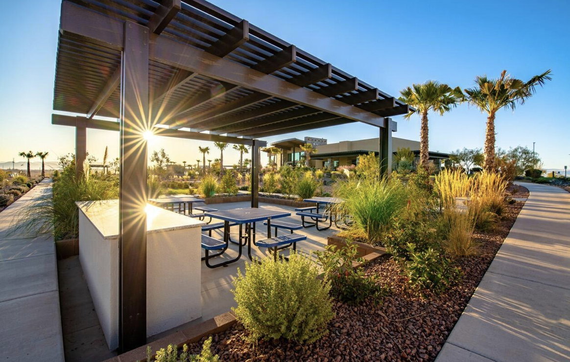 Outdoor covered patio featuring a built-in counter with a light-toned countertop, multiple picnic tables, and abundant landscaping with mature palm trees