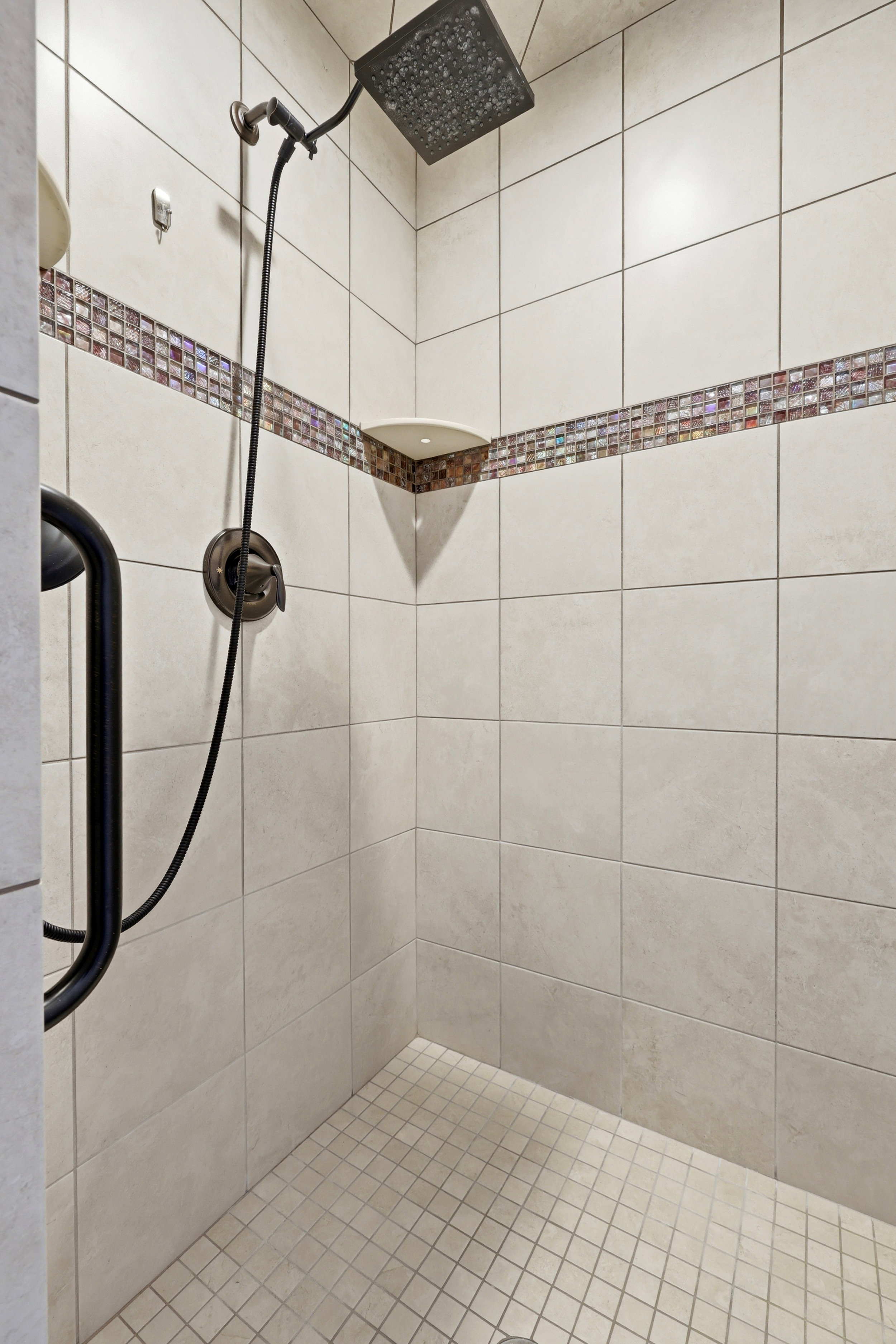 Master shower stall with square wall tiling, mosaic tile accent band, corner shelf, and rain showerhead with handheld sprayer