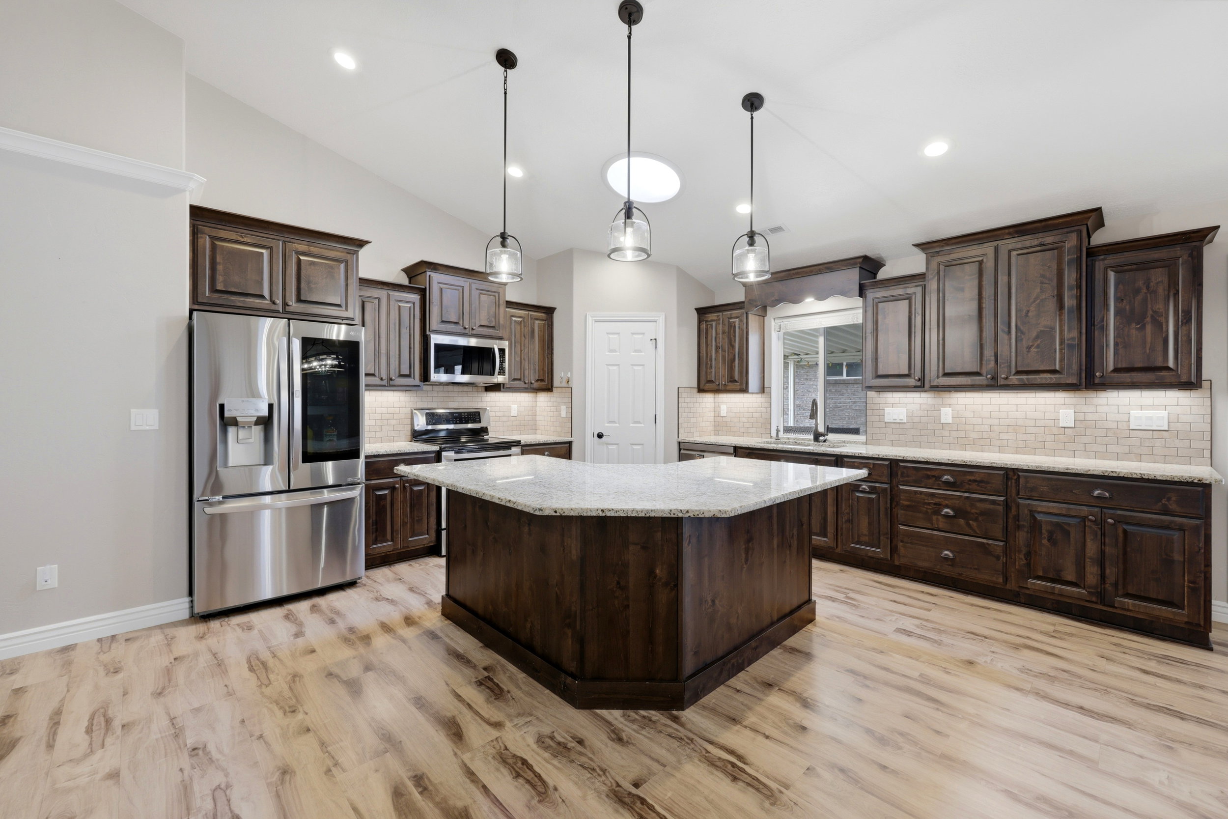Spacious kitchen featuring an angular island with a light-toned countertop, wood-finish flooring, dark wood cabinetry, a subway tile backsplash, and stainless steel appliances