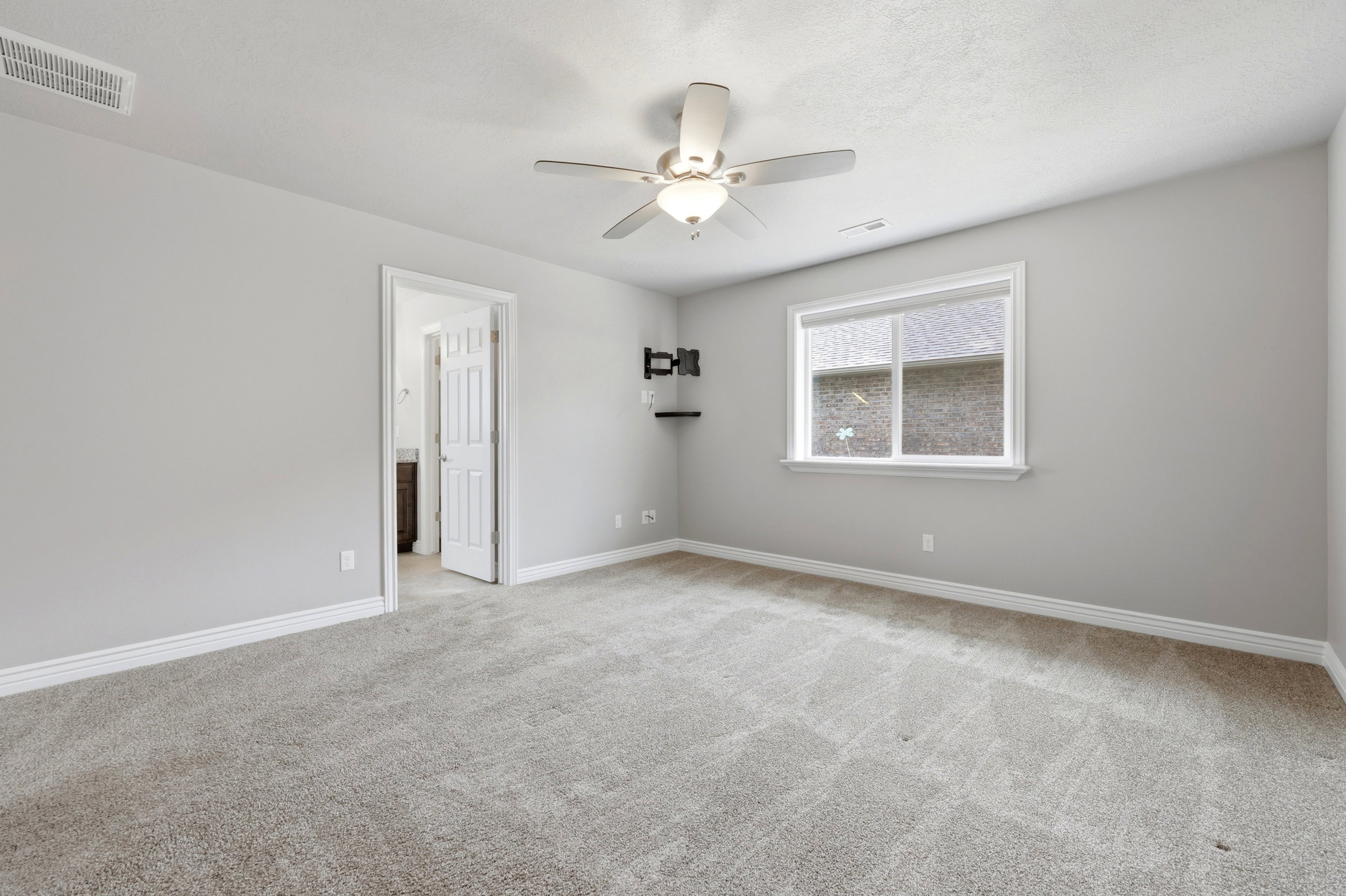 Spacious bedroom featuring light gray walls, plush carpeting, a ceiling fan with integrated lighting, a white-trimmed window, and a wall-mounted television bracket