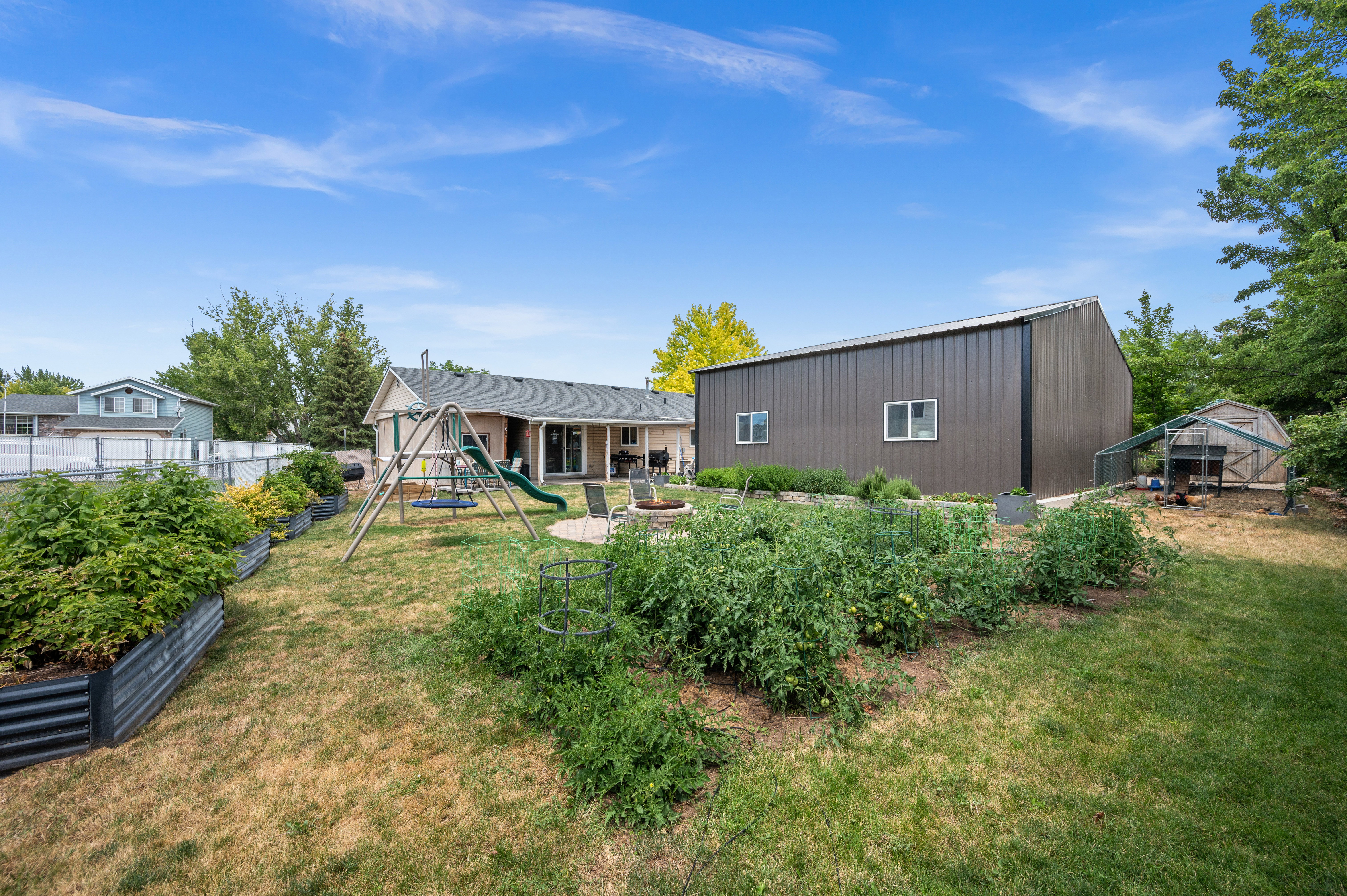 Spacious backyard featuring multiple garden beds, a large metal-sided outbuilding with windows, and a separate greenhouse structure