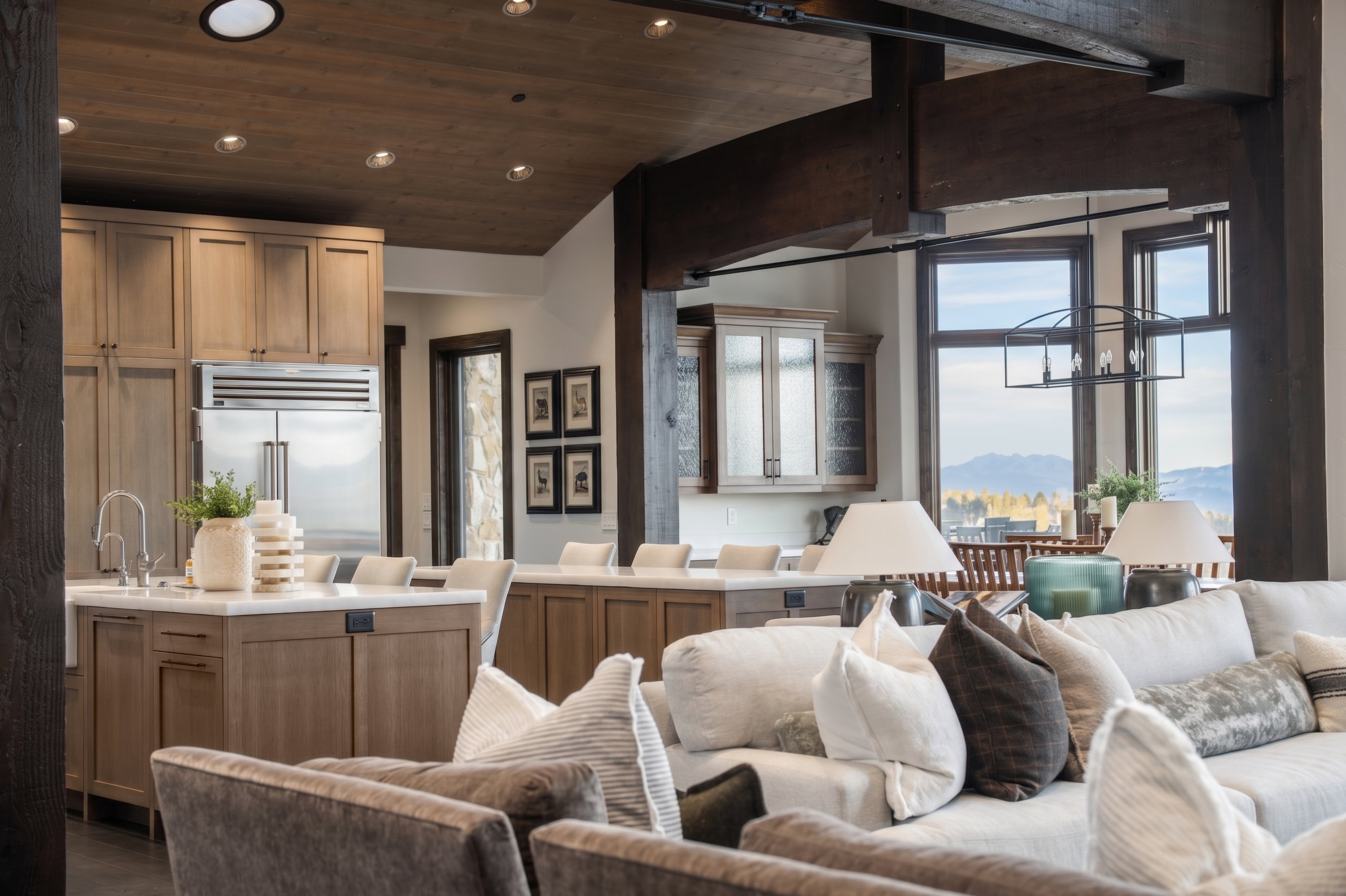 Open-concept interior featuring exposed wood beams, a kitchen island with seating, stainless steel refrigerator, recessed lighting, and large windows