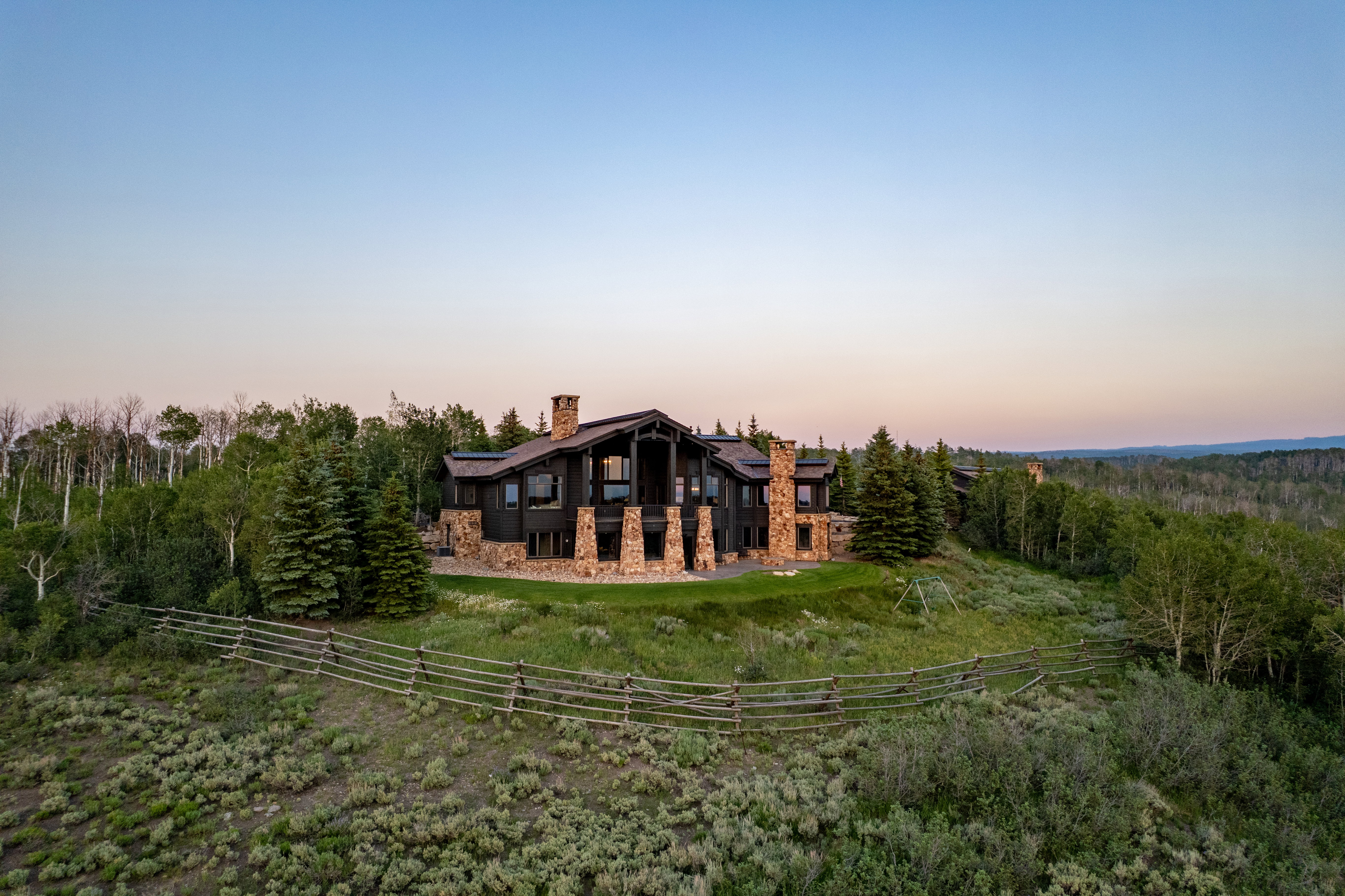 Expansive property featuring a multi-level structure, stone foundation and chimneys, dark siding, multiple windows, and natural landscaping with mature trees and a rustic fence
