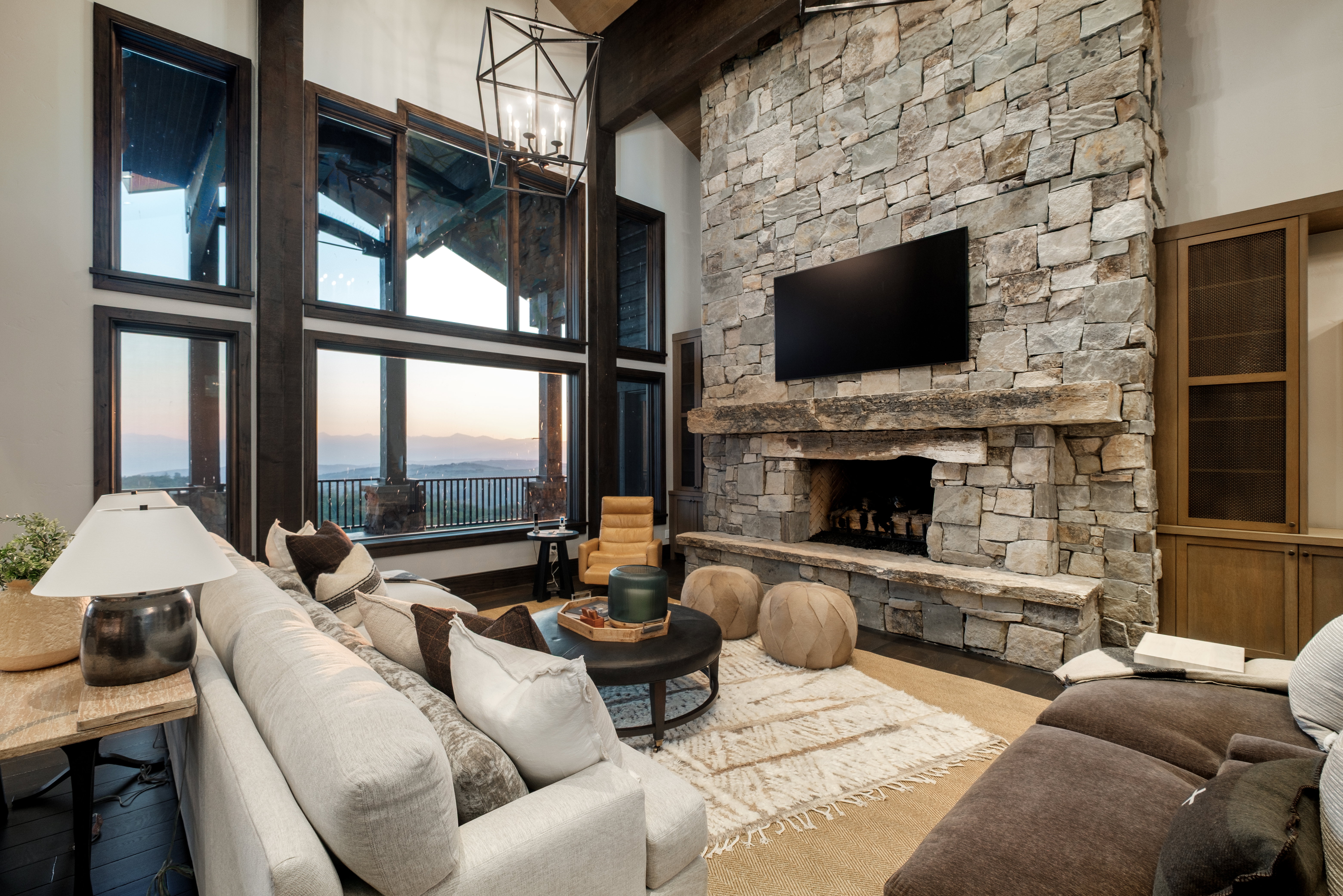 Floor-to-ceiling windows with dark wood framing, a stone-clad fireplace with a natural wood mantel, built-in wooden cabinetry, and exposed ceiling beams