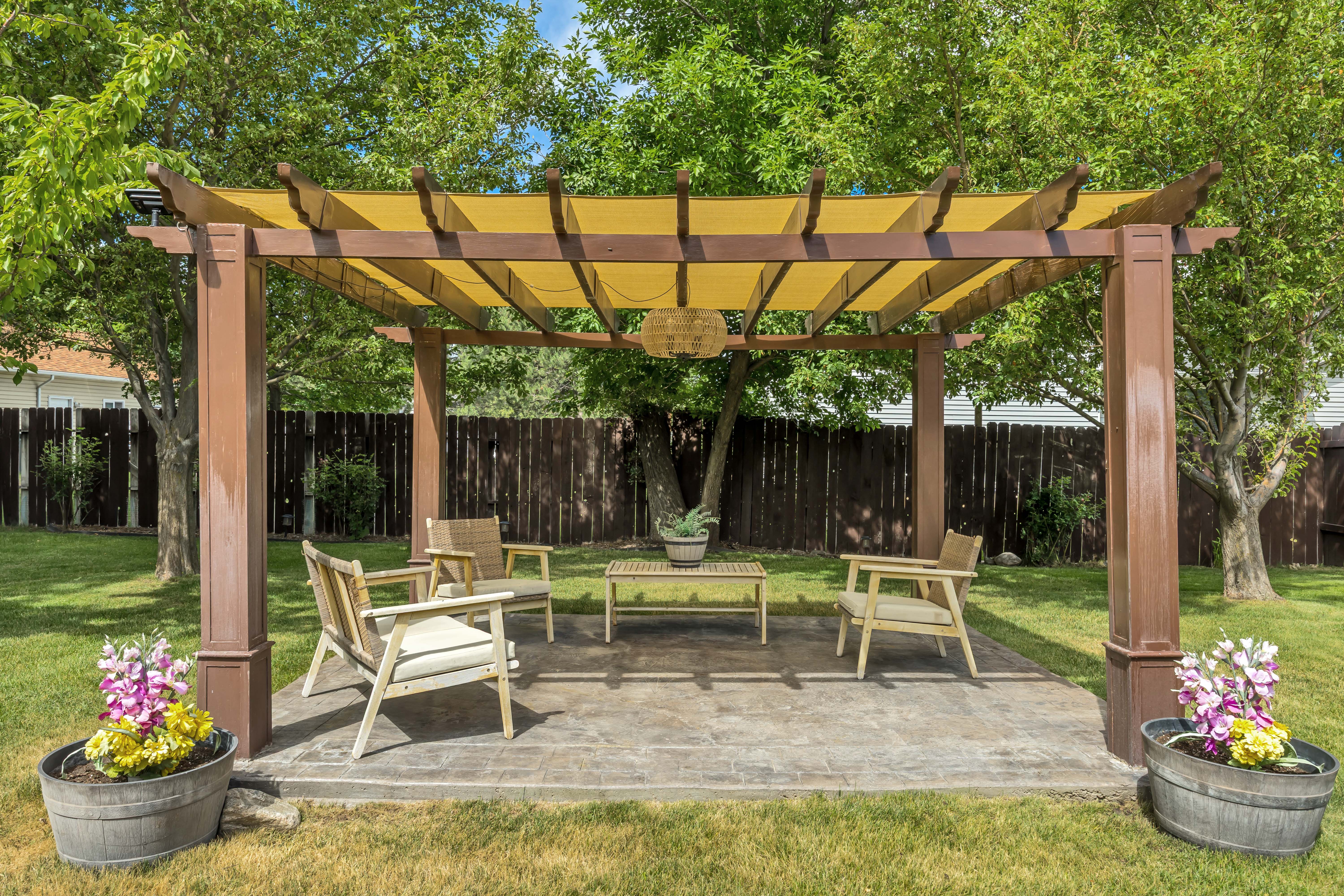 Expansive backyard featuring a pergola with a yellow fabric canopy, a stone paver patio, mature trees, and a wooden privacy fence