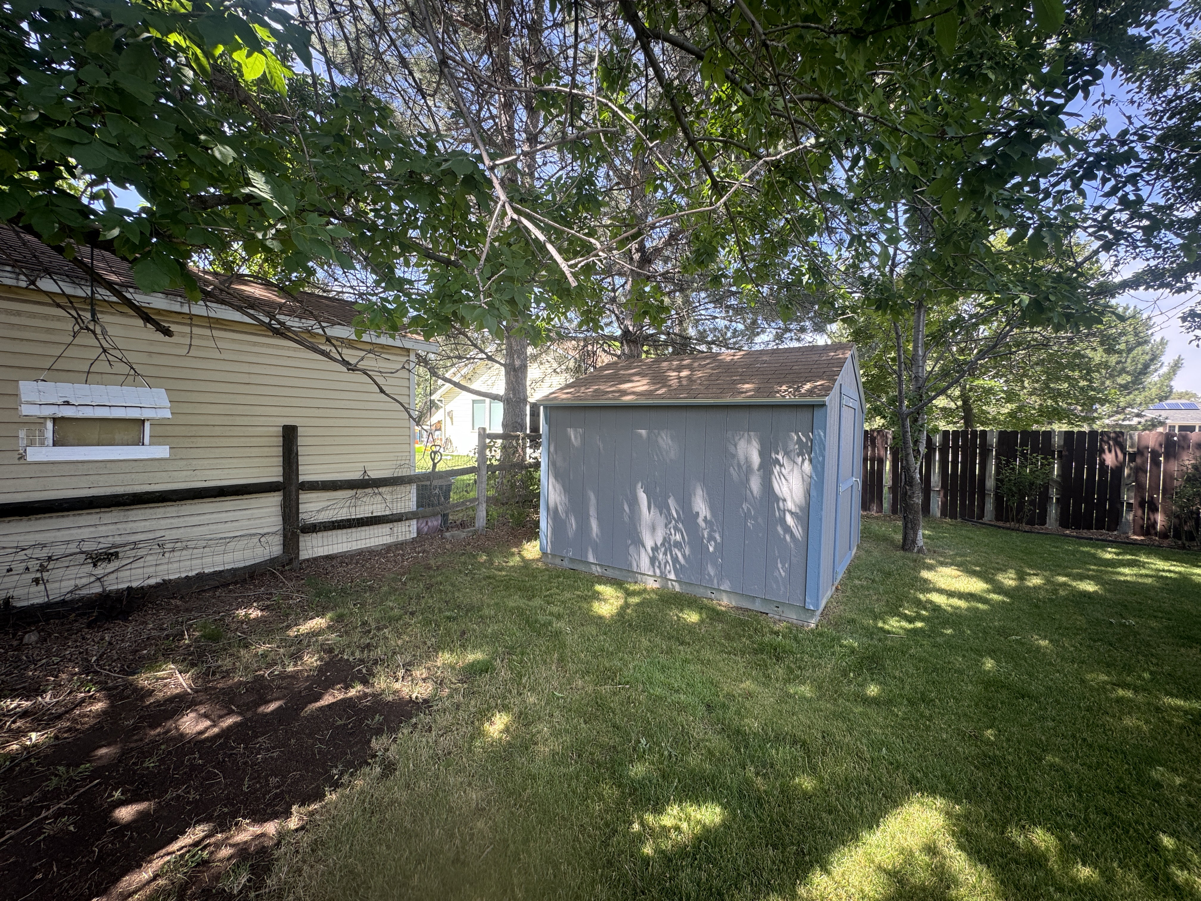 Backyard with a light blue storage shed featuring a brown shingled roof, a wooden privacy fence, mature trees, and a manicured lawn