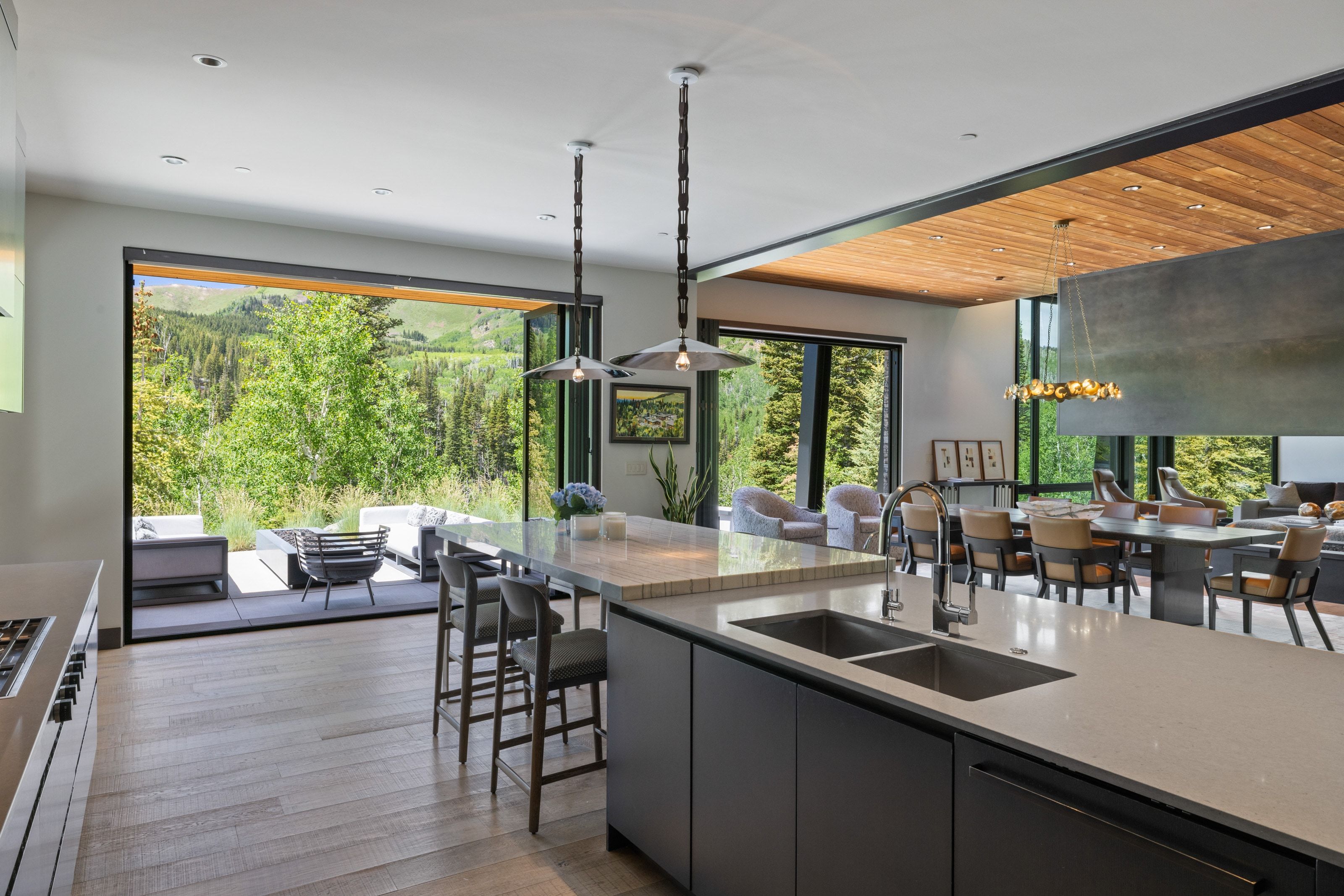 Open-concept kitchen featuring a large island with dual sinks, light wood-finish flooring, a wood-paneled ceiling, and a full-height glass wall opening to an outdoor patio with mountain scenery
