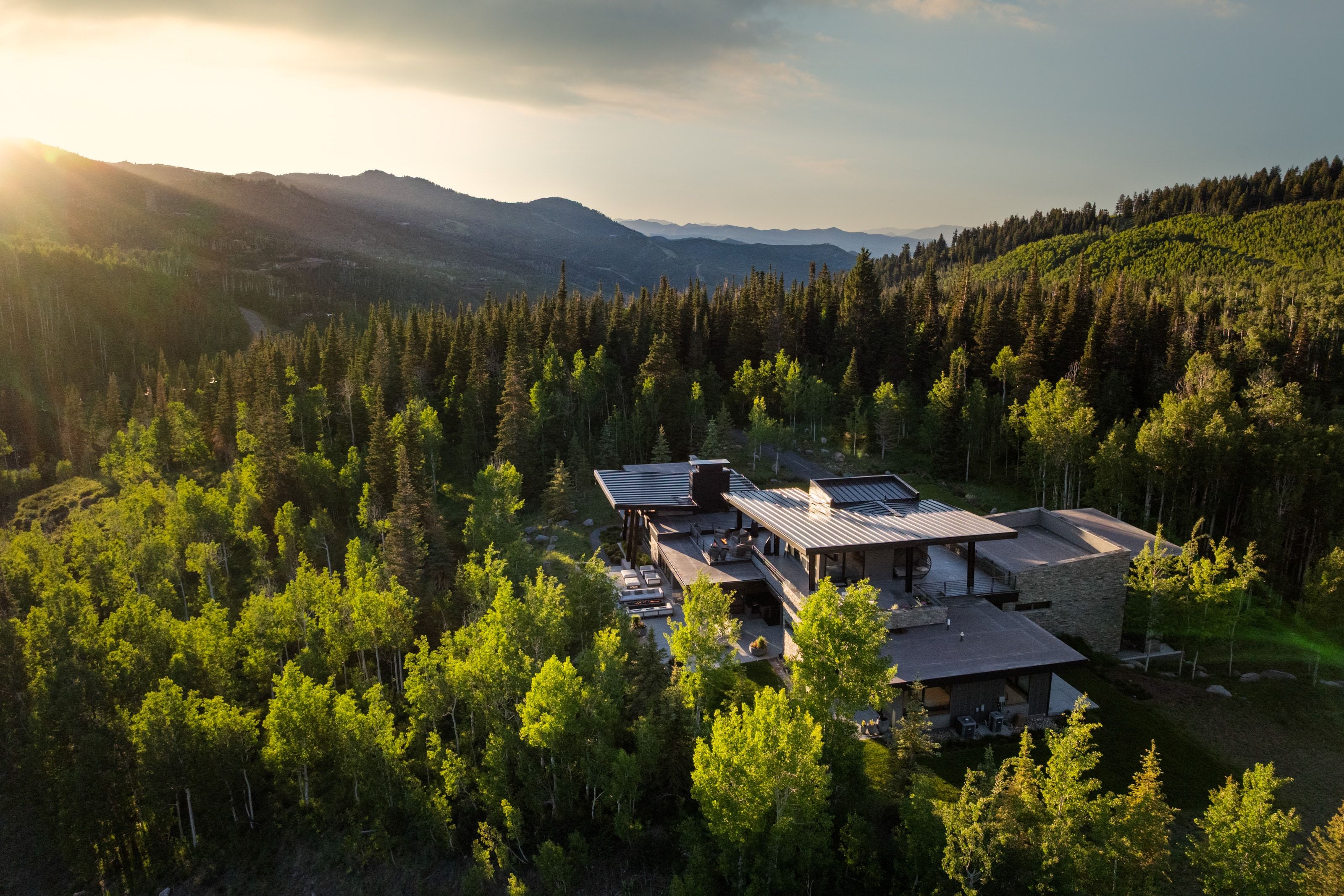 Expansive modern residence with a metal roof and multiple outdoor living areas, nestled among mature trees and mountains