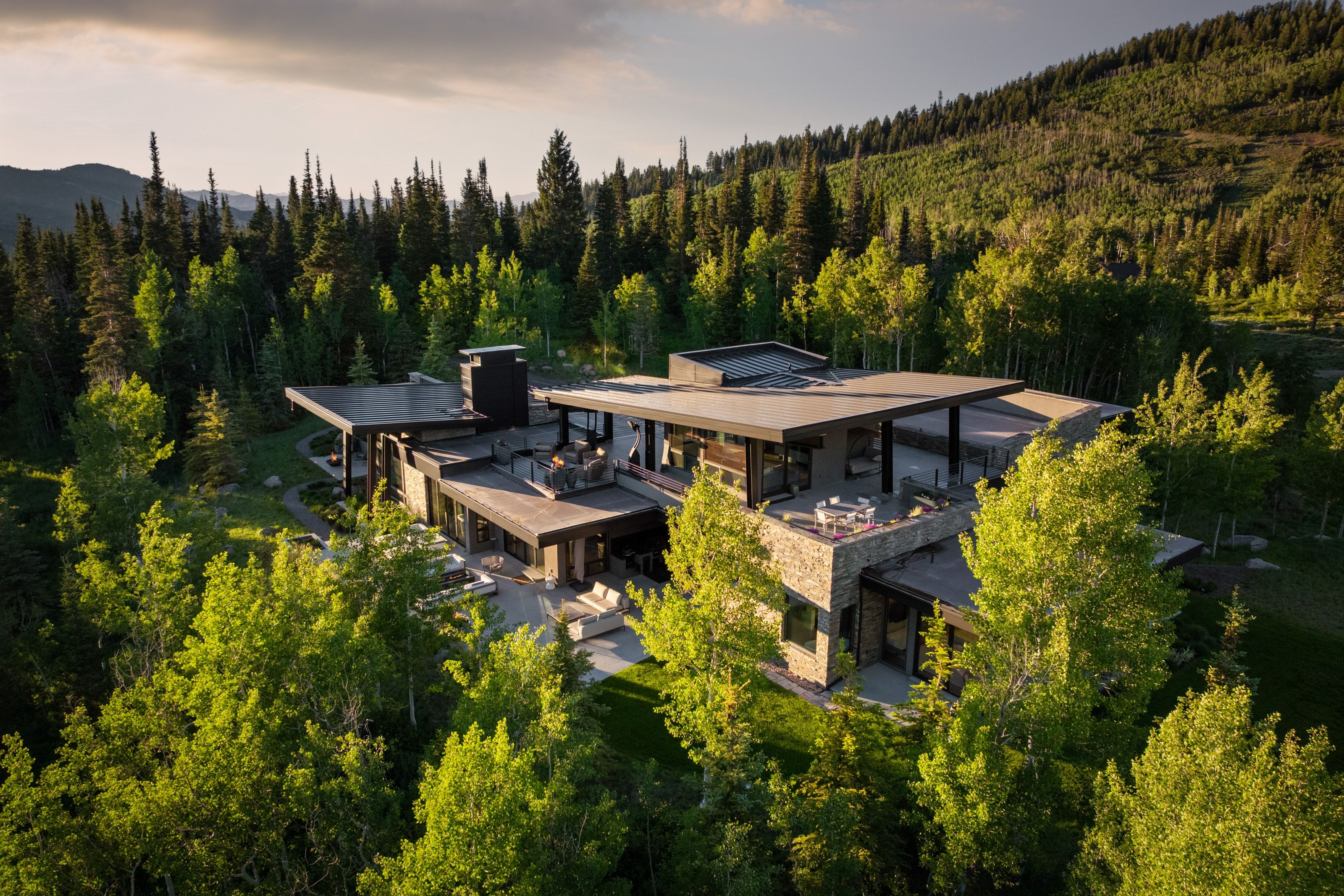 Contemporary mountain residence featuring stone and dark paneling, extensive flat rooflines, multiple outdoor patio areas, and a lush forested setting