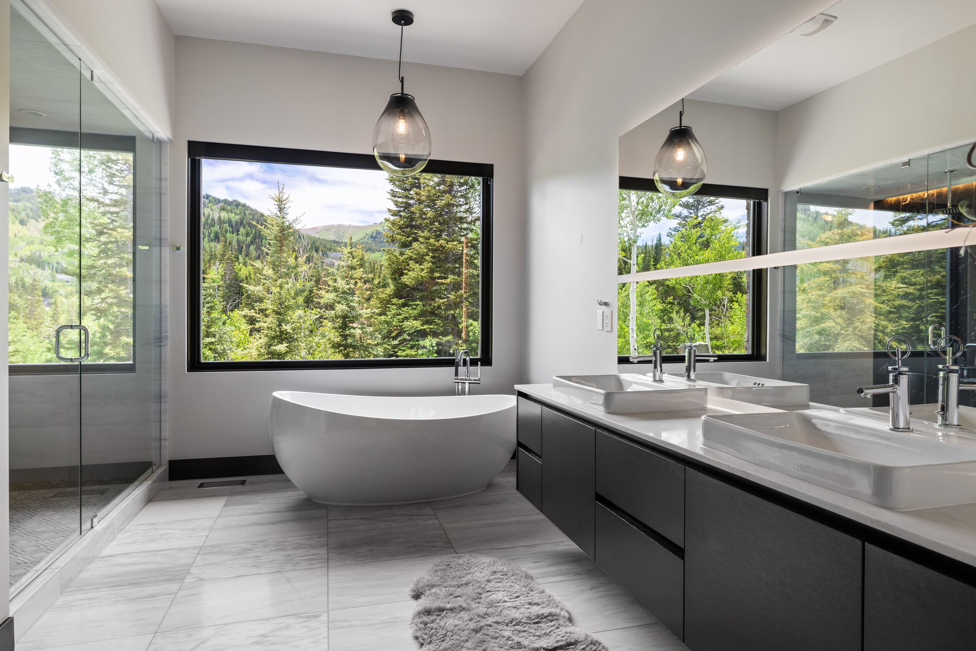 Modern bathroom featuring a freestanding soaking tub, expansive window with mountain scenery, dual vessel sinks atop a floating vanity, a large walk-in shower with glass enclosure, and light-toned tile flooring