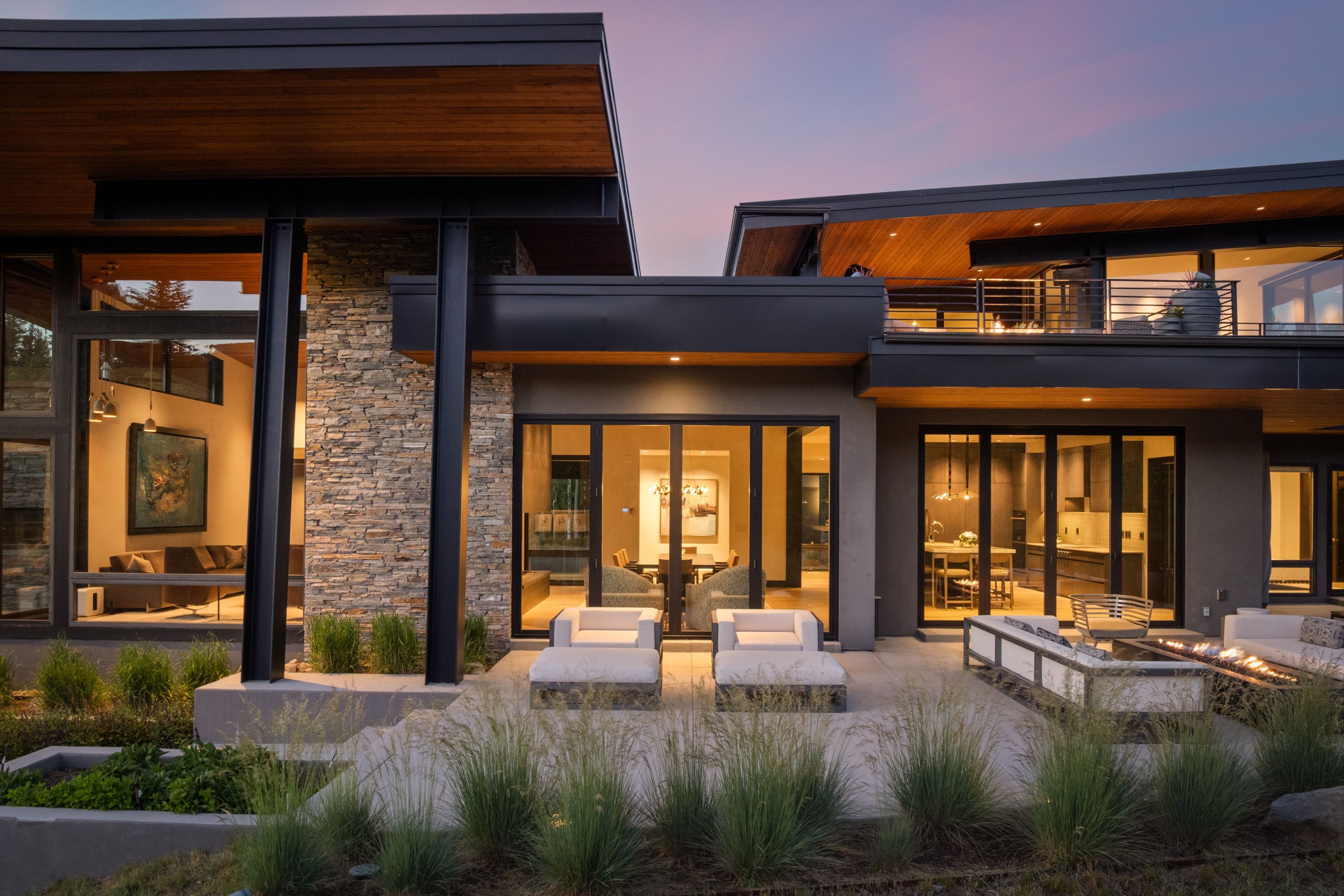Contemporary exterior featuring stone accents, expansive glass doors, multiple patios, wood-finish eaves, and a second-story balcony