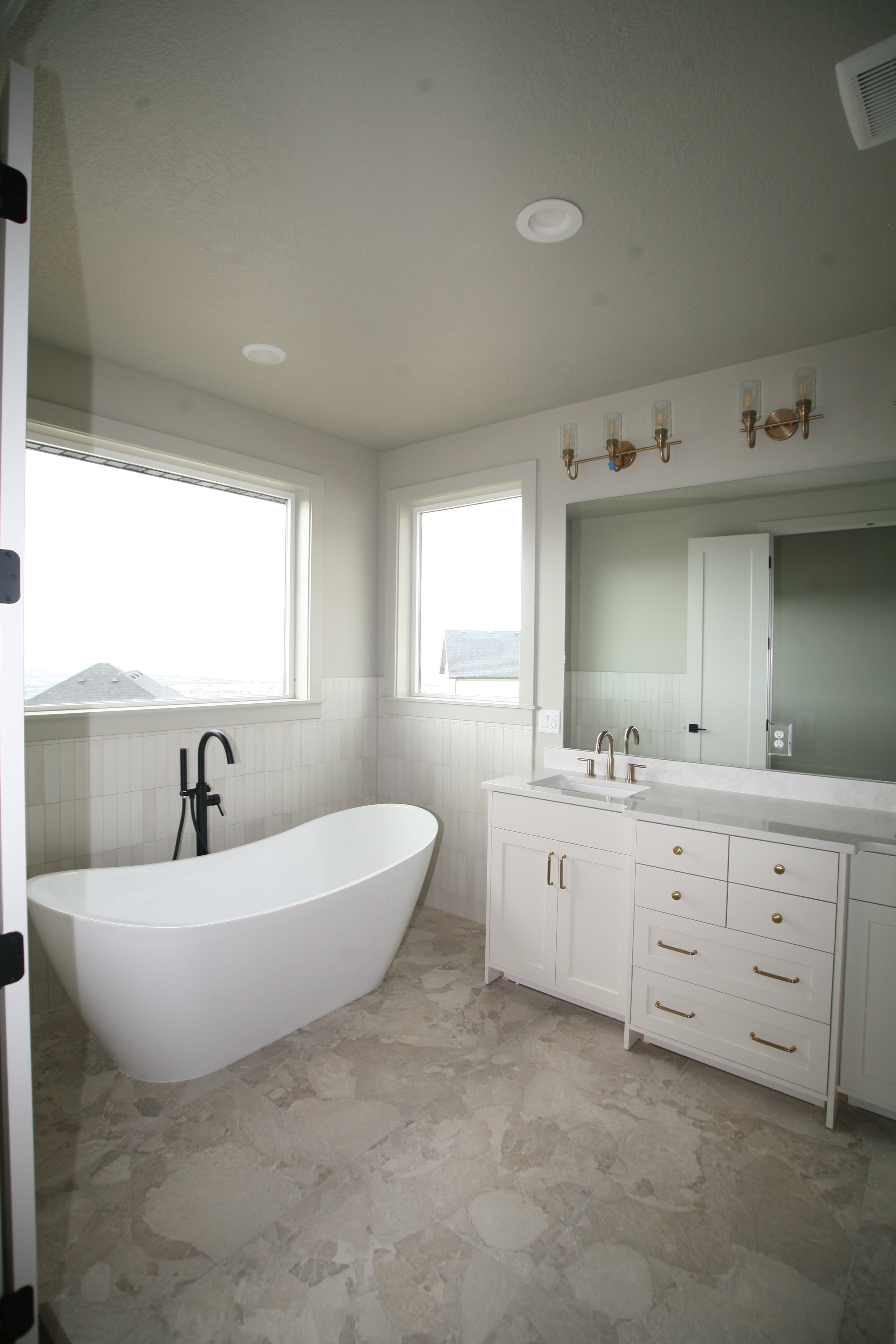 Soaking tub with a matte black floor-mounted faucet, white tile wainscoting, and large format stone-finish flooring