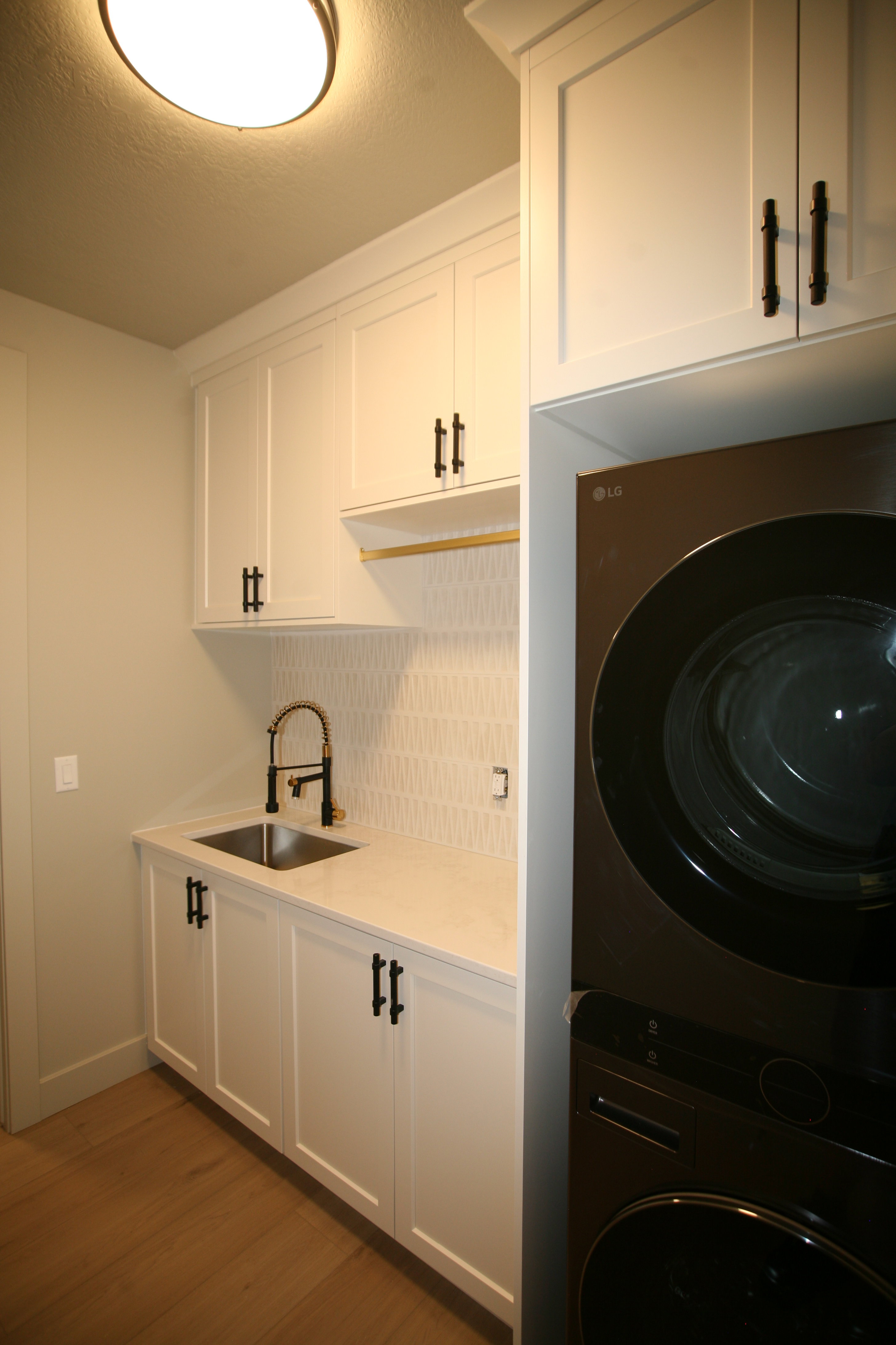 Laundry room featuring white shaker-style cabinetry with matte black hardware, a stainless steel sink with a matte black pull-down faucet, light-toned wood-finish flooring, and a stacked washer and dryer unit
