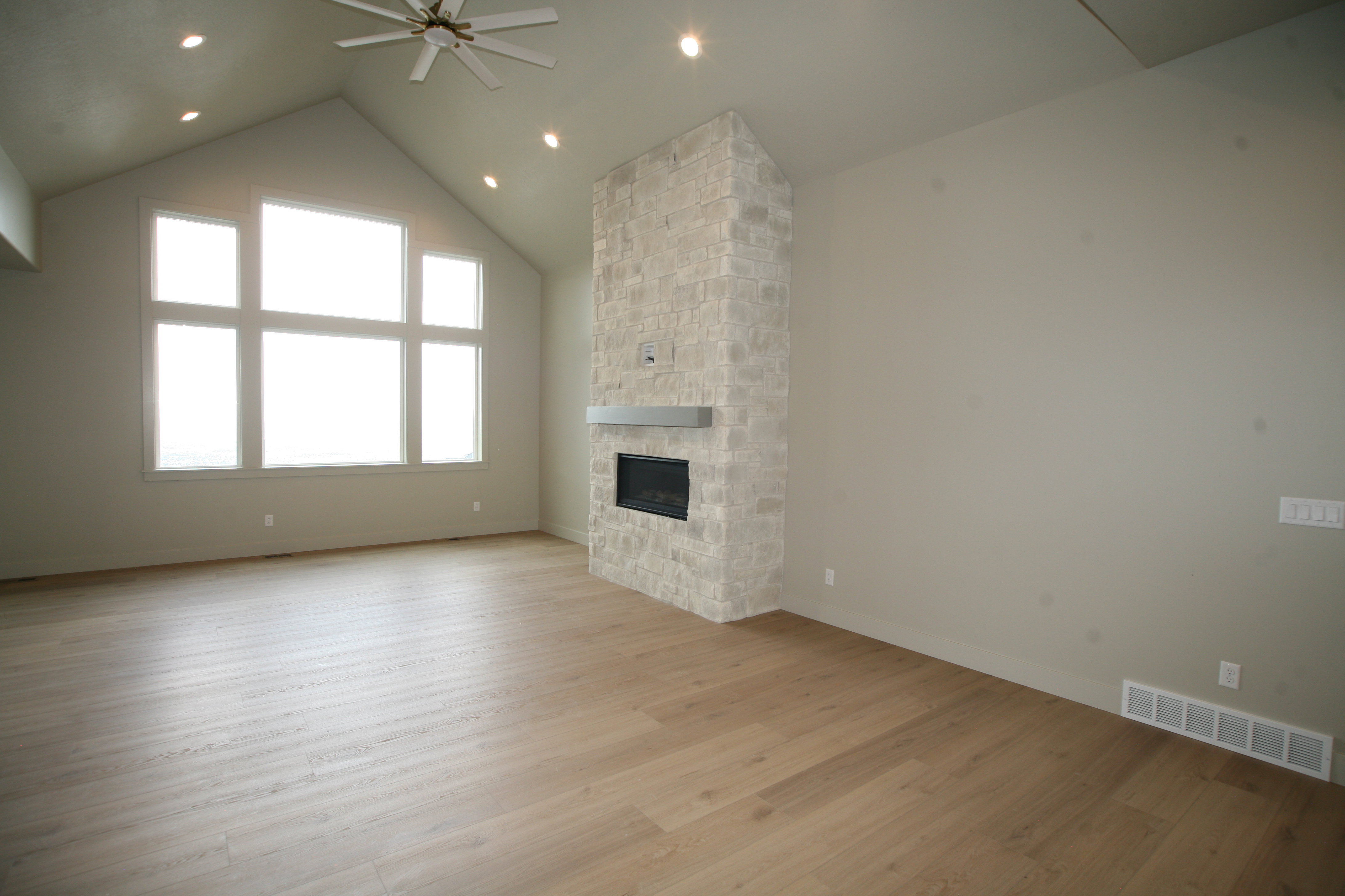 Spacious room featuring vaulted ceilings, a large multi-pane window array, a stacked stone fireplace with a floating mantel, recessed lighting, and wood-finish flooring