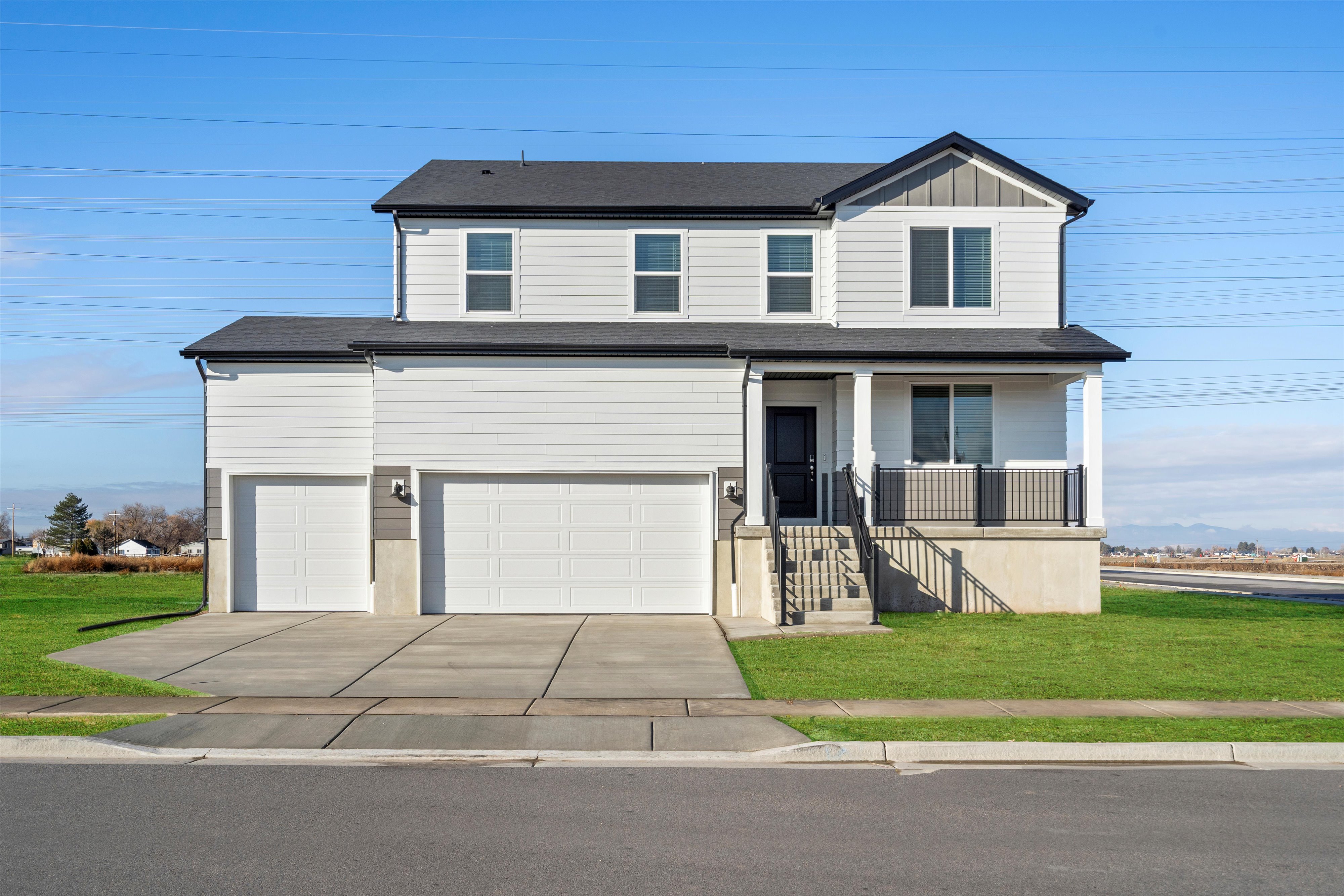 Two-story residence featuring light-colored siding with dark trim accents, a covered front porch with dark railings, a dual garage, a concrete driveway, and a front lawn