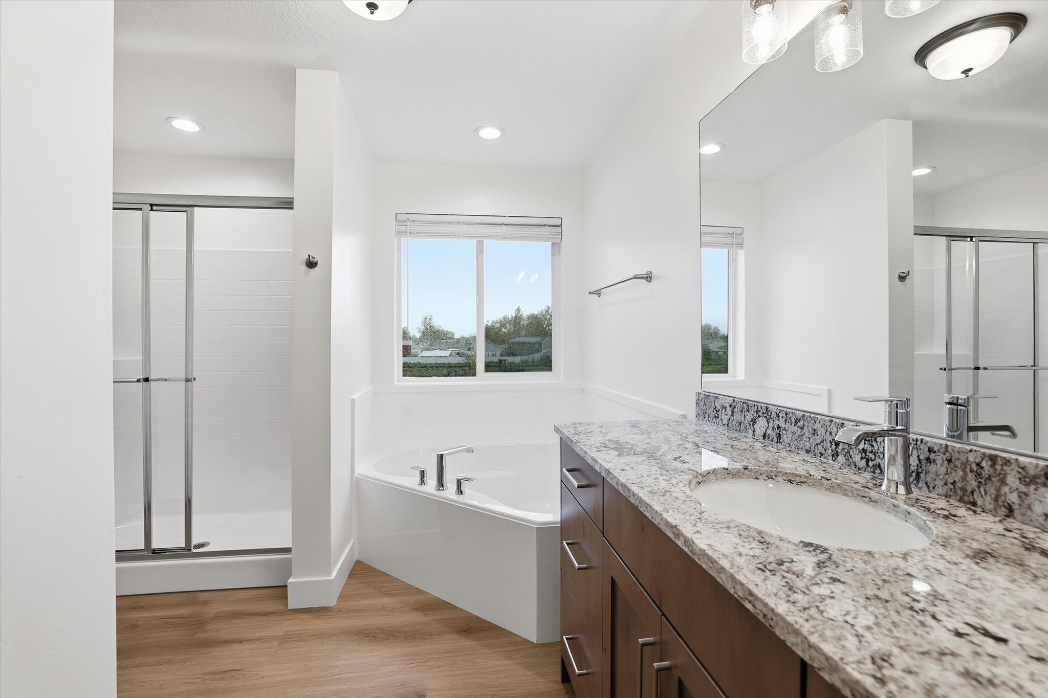 Spacious bathroom featuring a double vanity with stone countertops, a large soaker tub under a window, a separate shower with glass enclosure, and wood-finish flooring