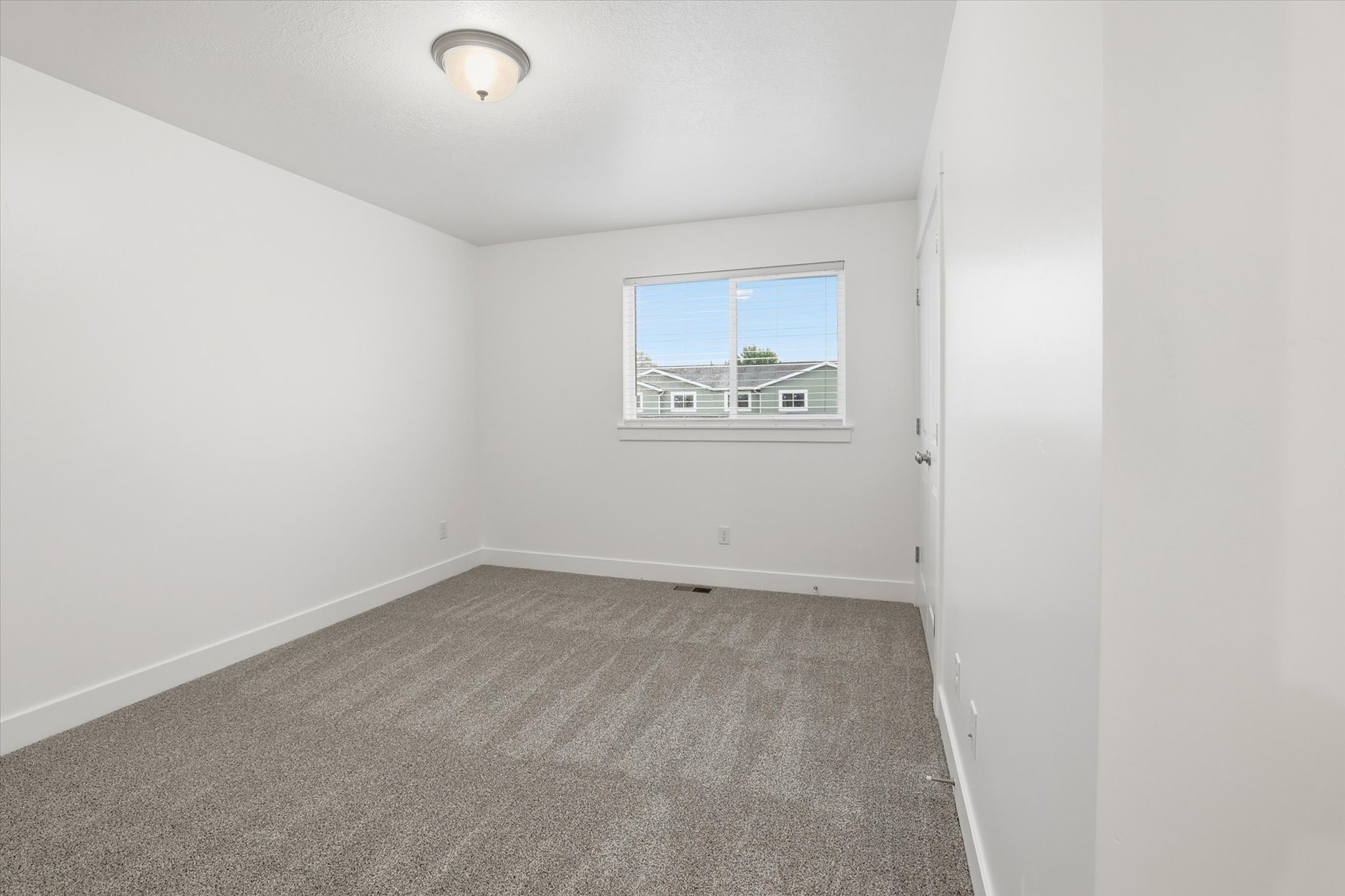 Neutral-toned room featuring a single window with blinds, light gray patterned carpeting, white baseboards, and a semi-flush mount ceiling light fixture