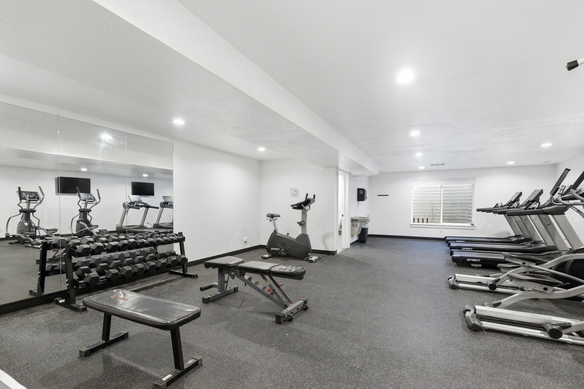Dedicated fitness area featuring rubberized flooring, mirrored wall, recessed lighting, and a horizontal window with blinds