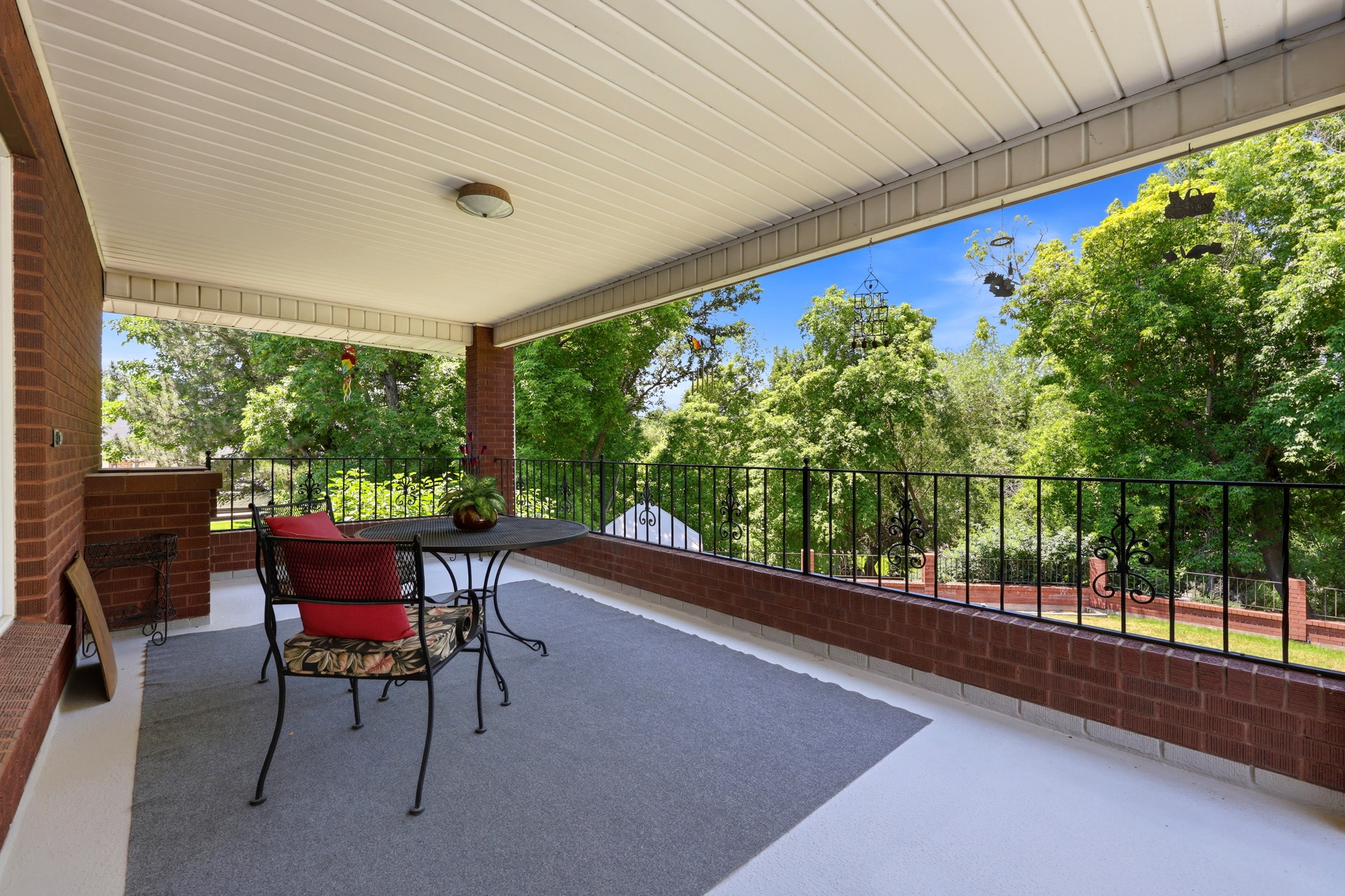 Covered outdoor patio featuring brick walls, an ornamental metal railing, and a slatted ceiling with a flush-mount light fixture