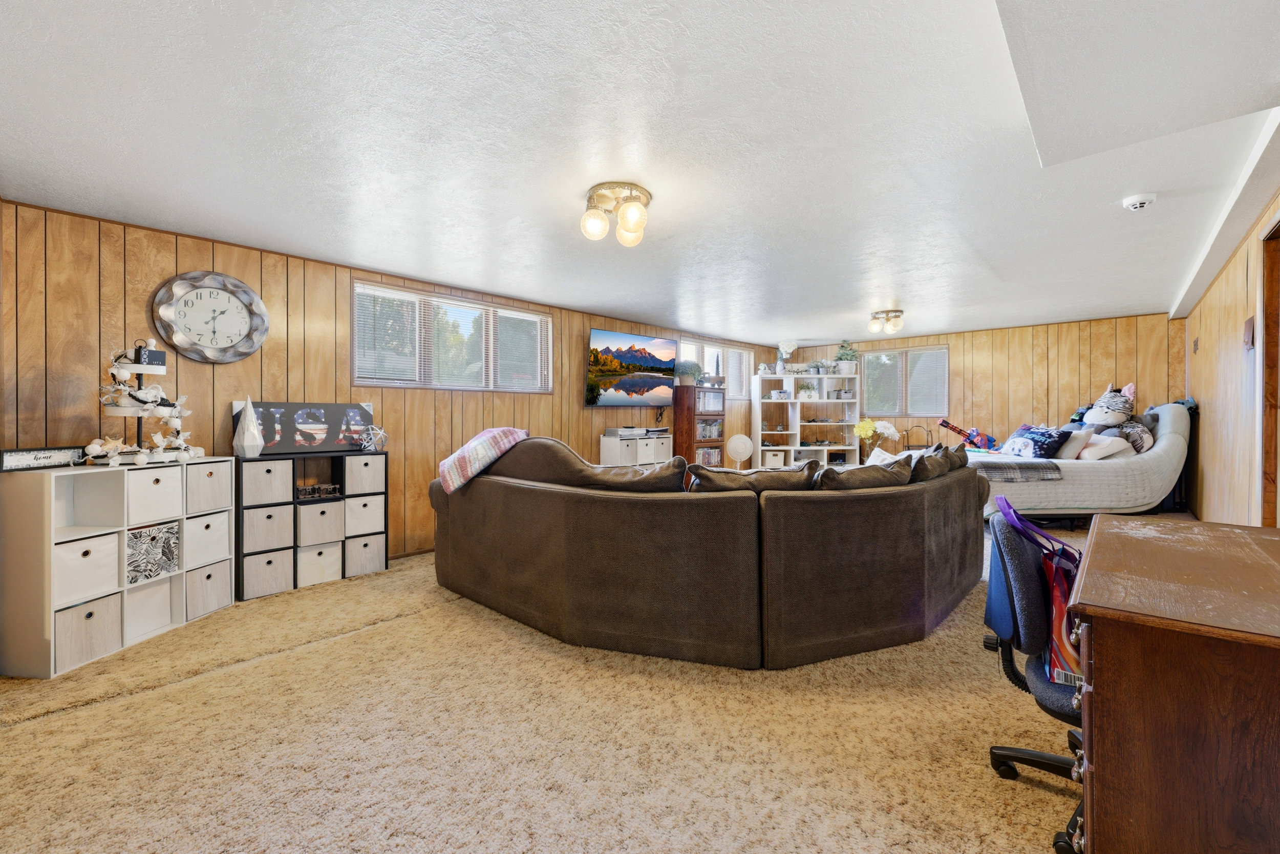 Spacious lower-level room featuring wood paneling, light-colored carpeting, multiple windows, and overhead lighting fixtures