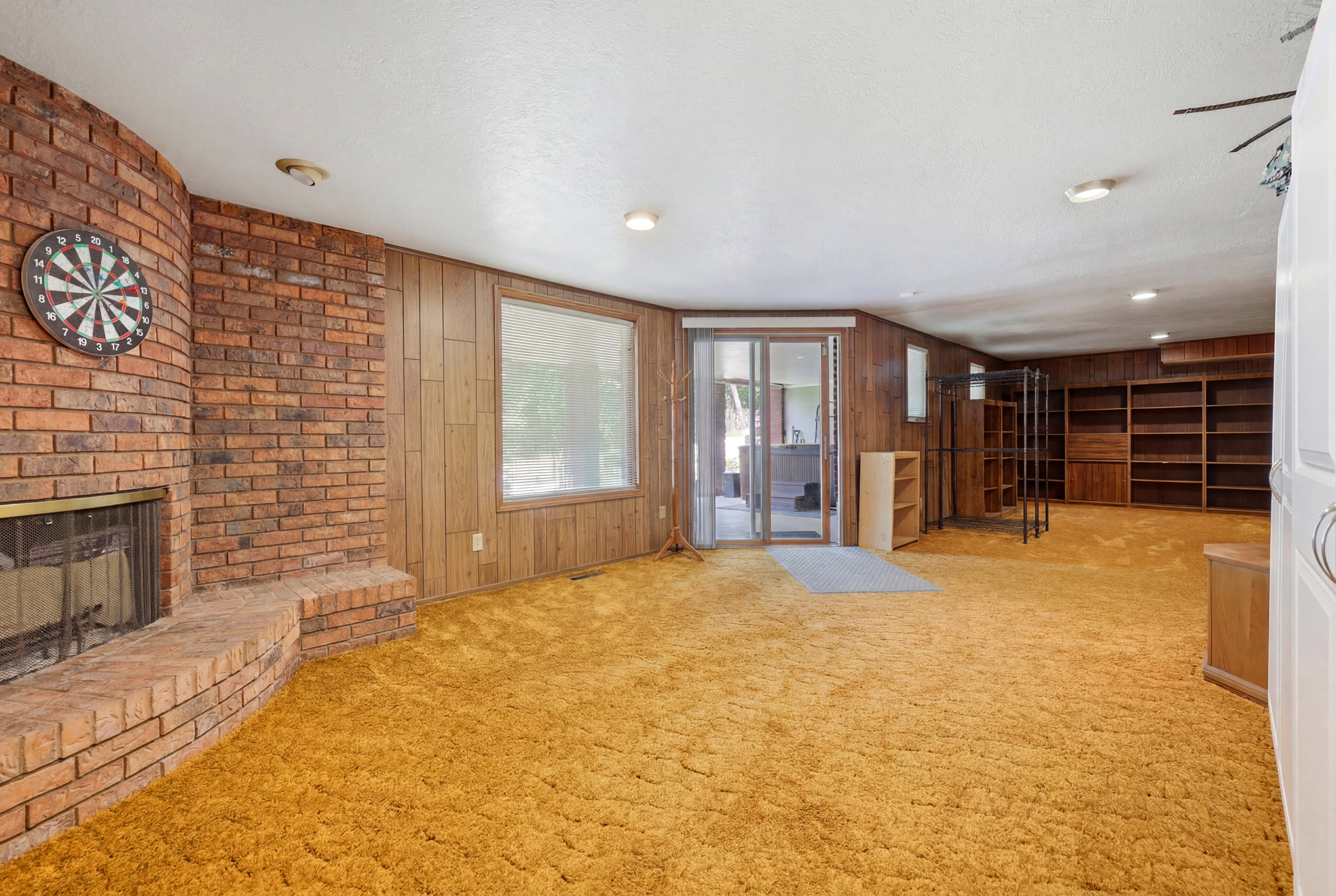 Spacious room featuring a brick fireplace with hearth, wood-paneled walls, built-in shelving, and a large window