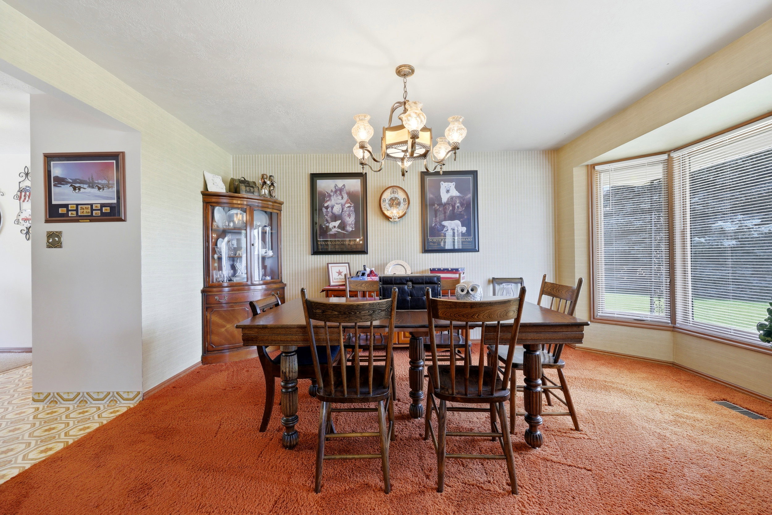 Spacious dining area featuring a prominent bay window, patterned wallpaper, overhead chandelier lighting, and direct access to an adjacent room with patterned flooring