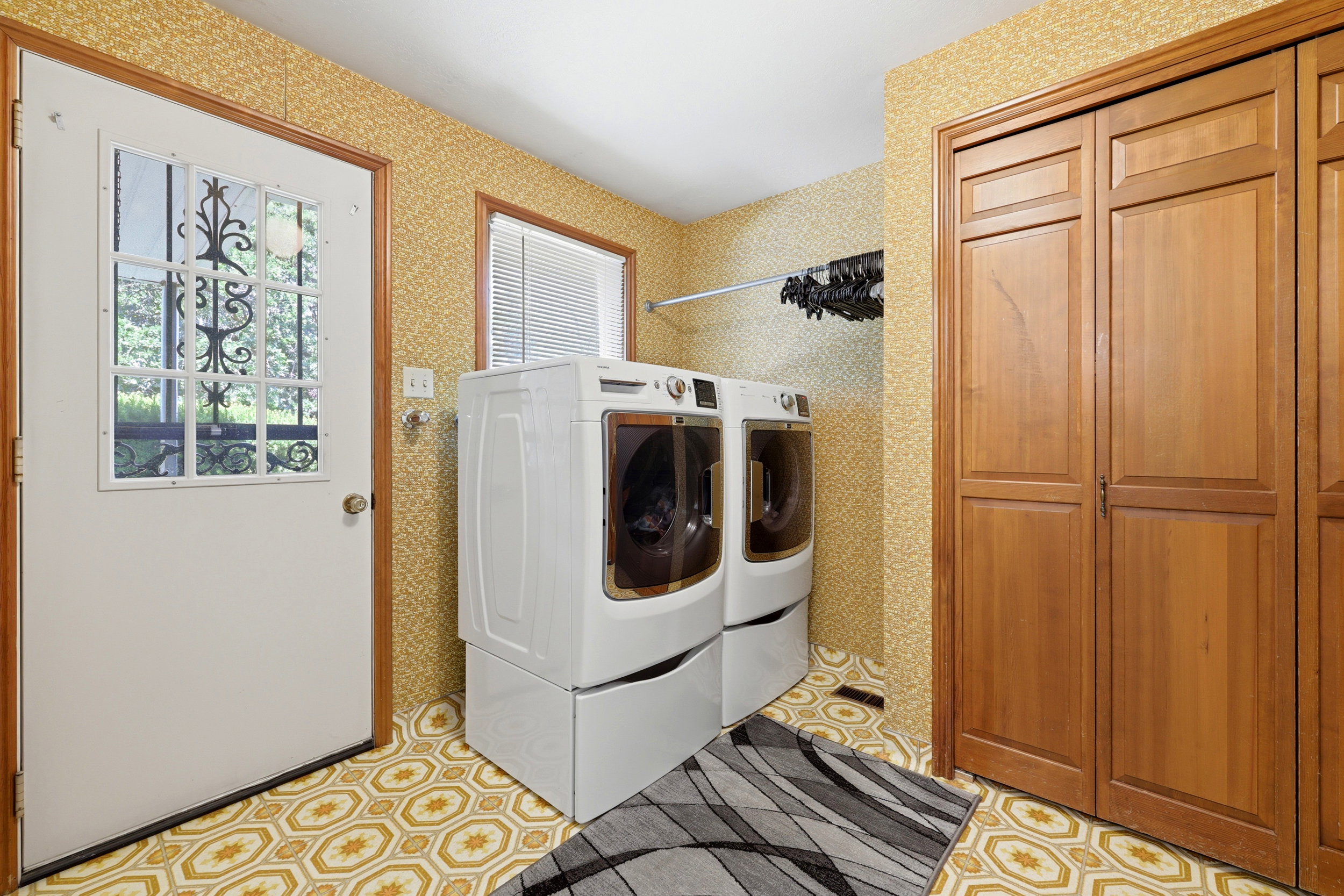 Utility area featuring patterned flooring, built-in wooden cabinetry, a window with blinds, and a door with decorative grilles
