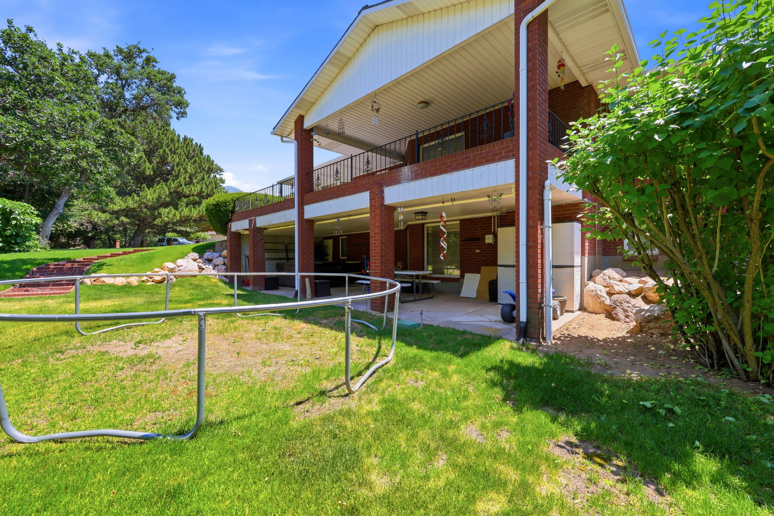 Expansive brick exterior featuring a covered patio, upper-level balcony with metal railing, and a sloped lawn with mature trees