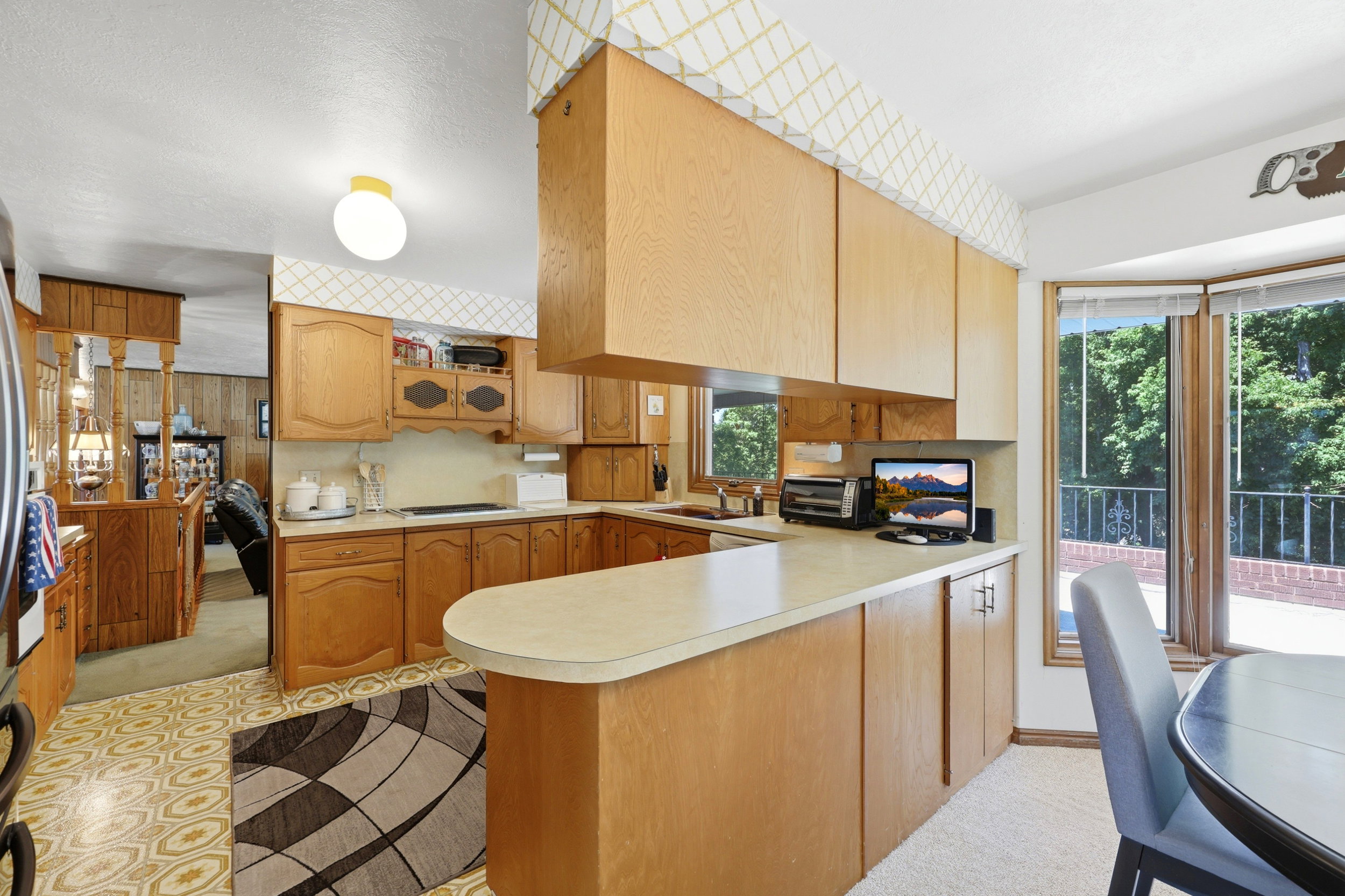 Kitchen featuring light wood cabinetry, a peninsula counter with a rounded edge, patterned floor tile, and a bay window with a surrounding outdoor brick patio