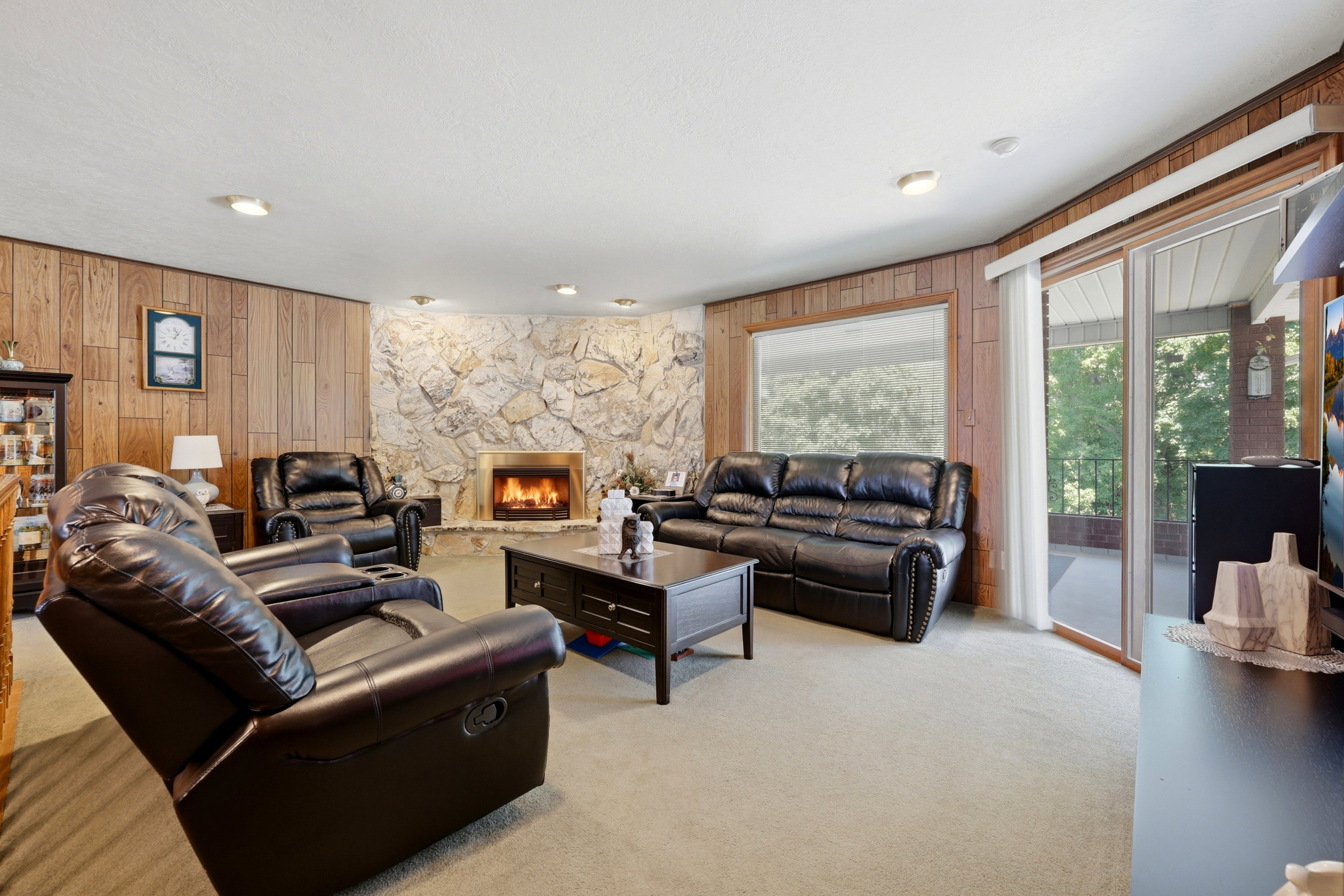 Living area featuring a stone-clad fireplace with brass surround, wood-paneled walls, light-toned carpeting, and a sliding glass door leading to an outdoor space