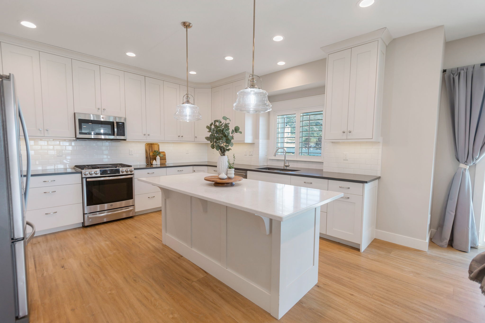 Expansive kitchen featuring a central island with a white countertop, white shaker-style cabinetry, stainless steel appliances, a white subway tile backsplash, and wood-finish flooring