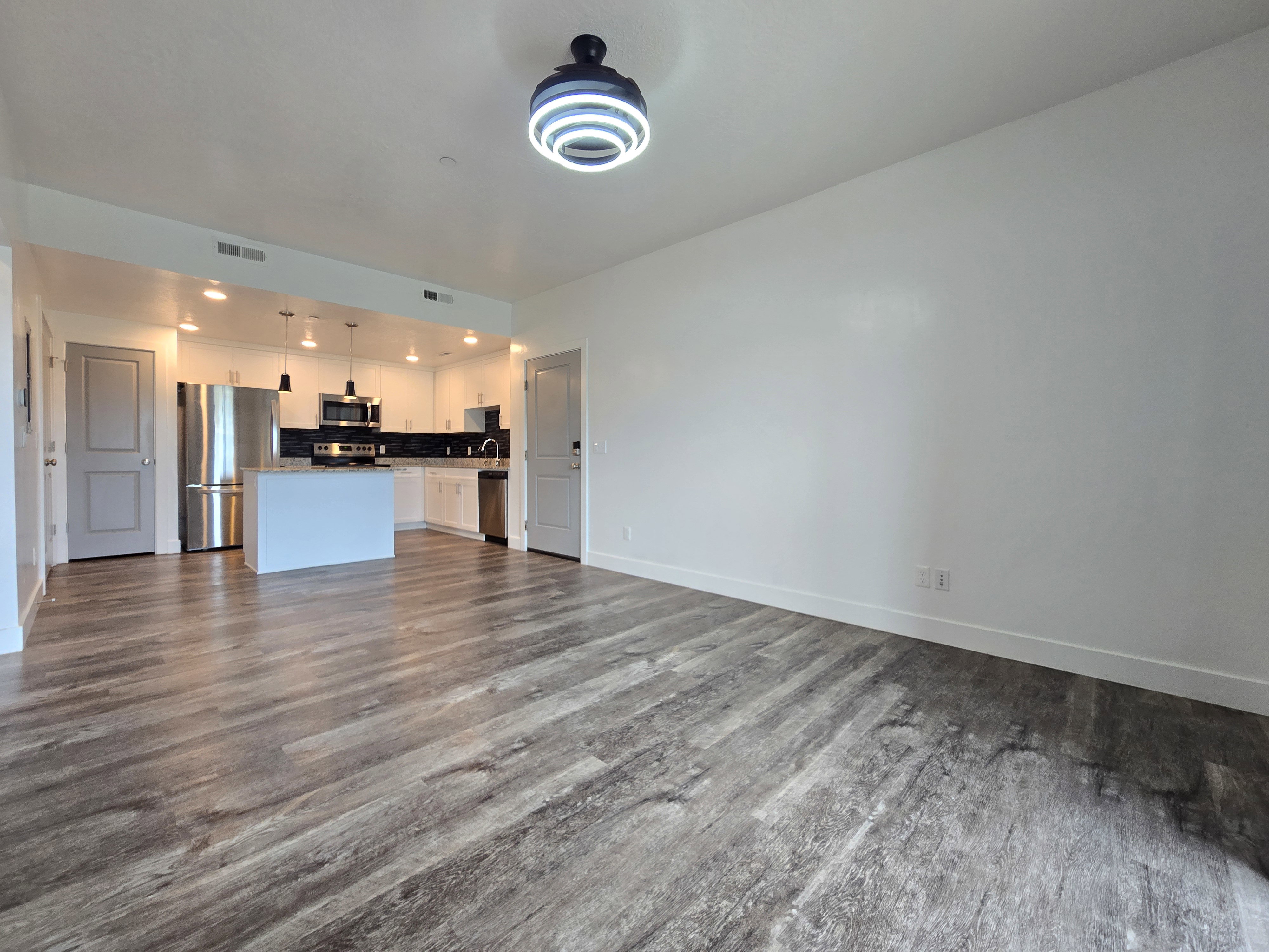 Open-concept living area featuring wood-finish flooring, a contemporary ceiling light fixture, and a kitchen with white cabinetry, stainless steel appliances, and an island