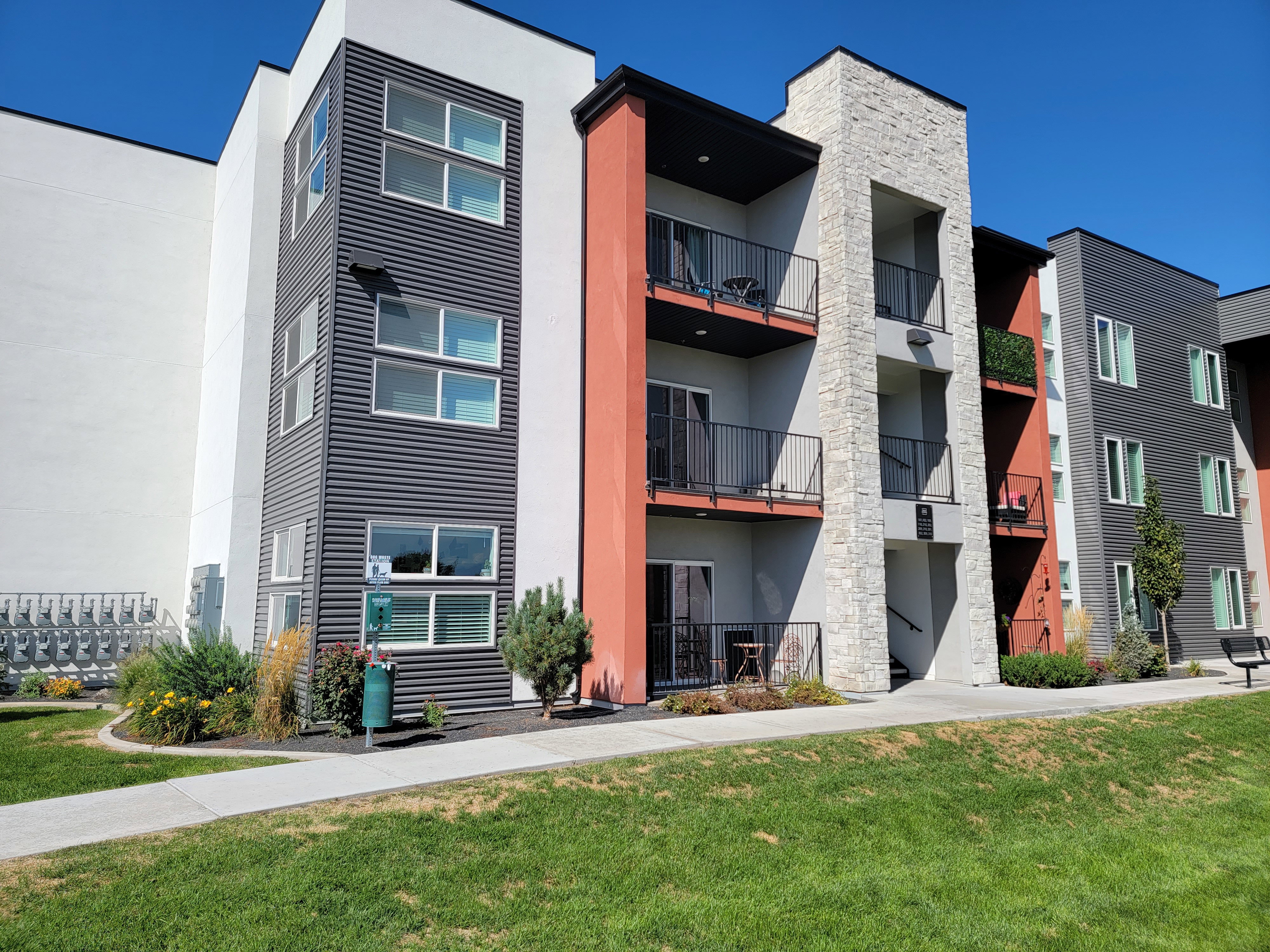 Contemporary multi-story facade featuring stone accents, private balconies with black railings, and a mix of white, red-orange, and dark gray siding