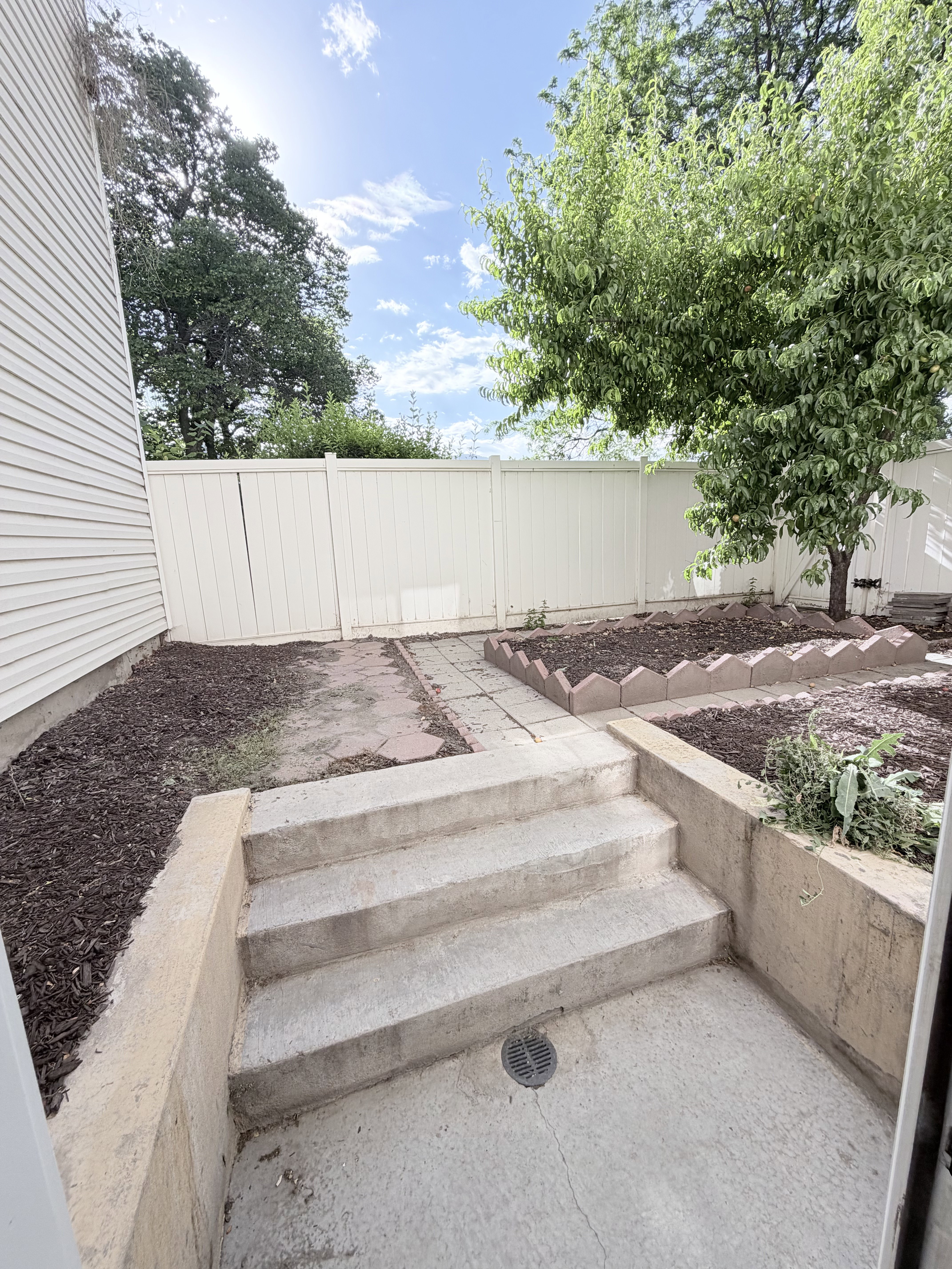 Concrete steps leading to a paved patio, featuring a privacy fence, raised garden beds, mature trees, and landscaping with mulch