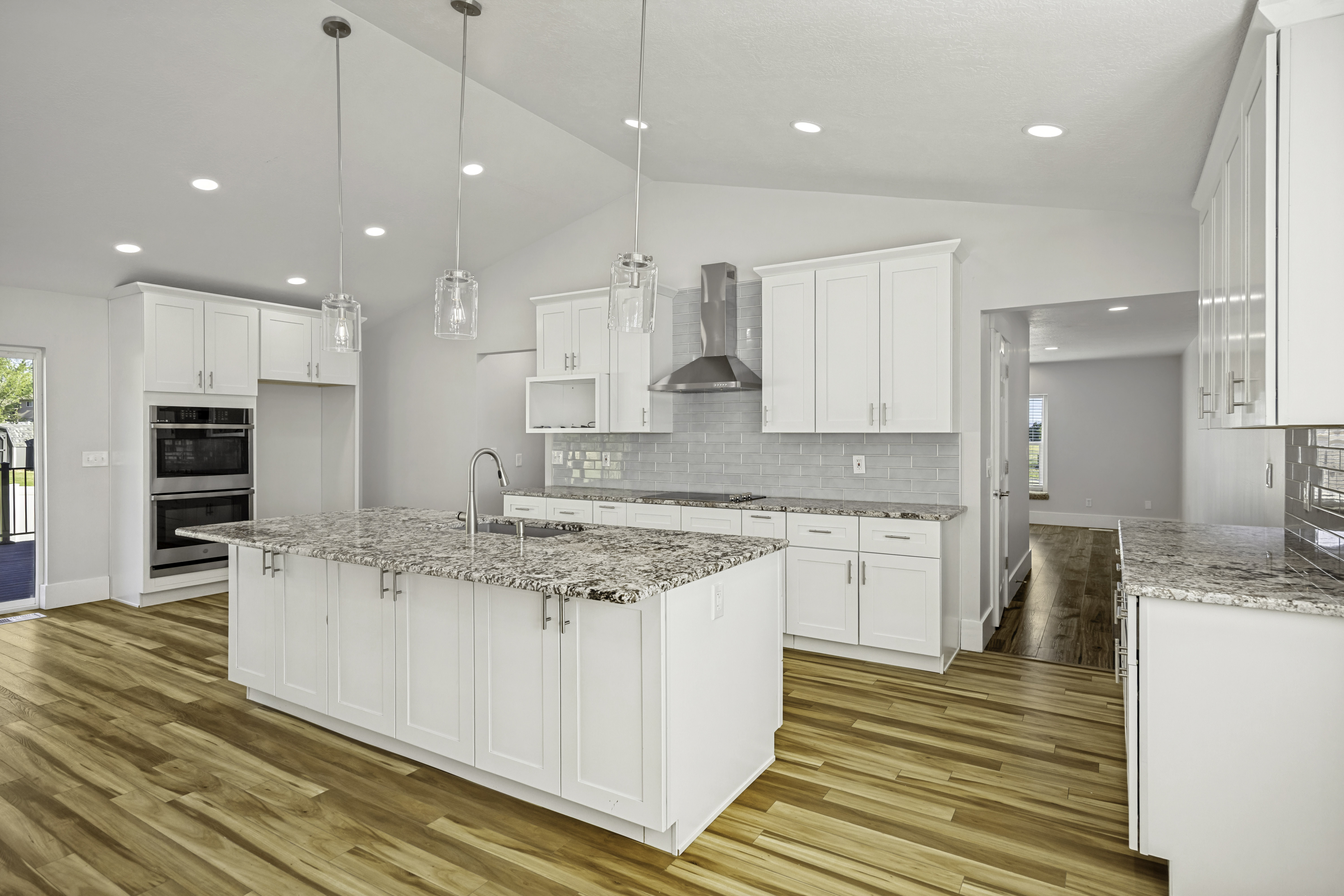 Open-concept kitchen featuring an expansive center island with an undermount sink, granite countertops, white shaker cabinetry, stainless steel double wall ovens, and wood-finish flooring