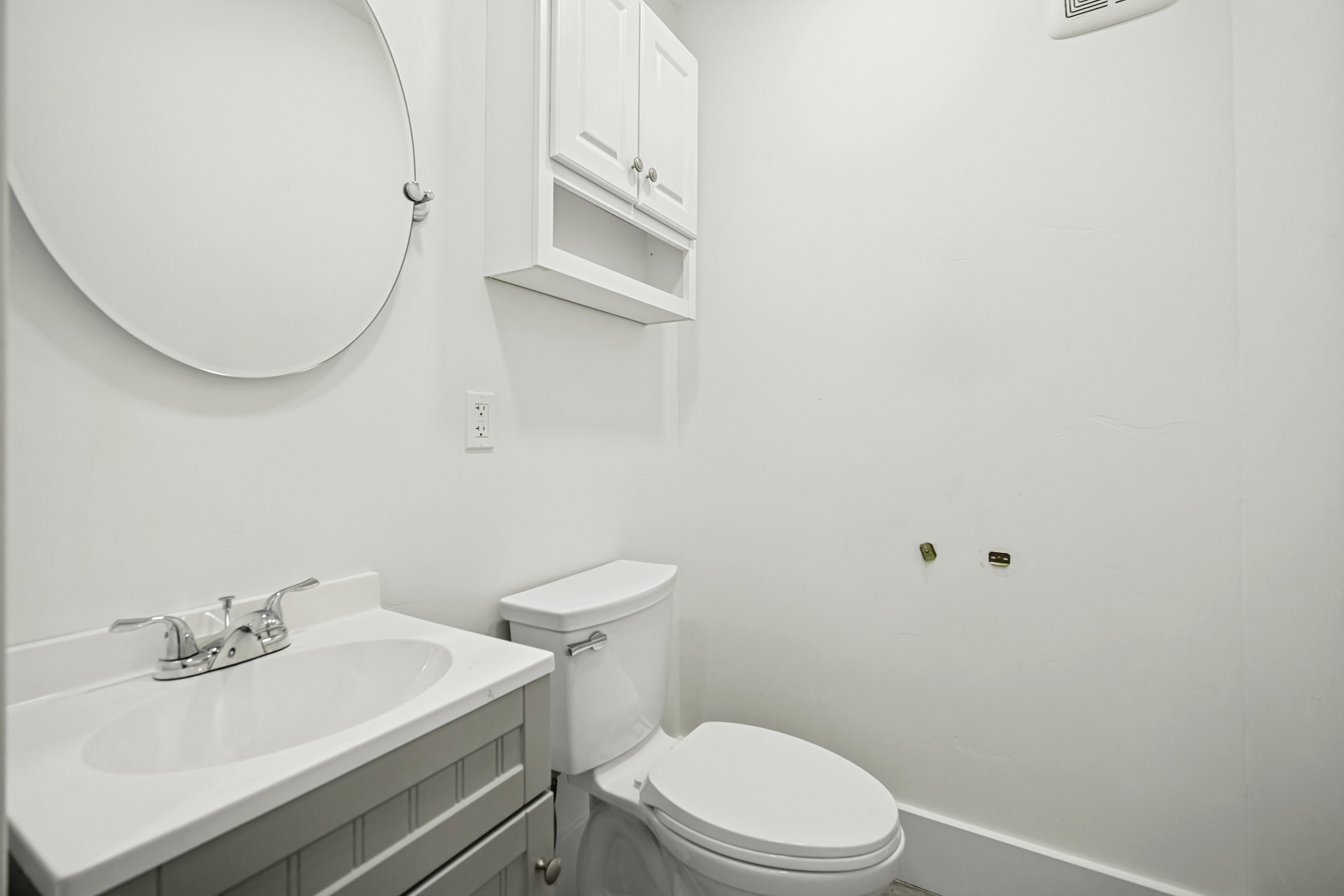 White-painted vanity with integrated sink and chrome faucet, round wall mirror, and white wall cabinet with open shelving