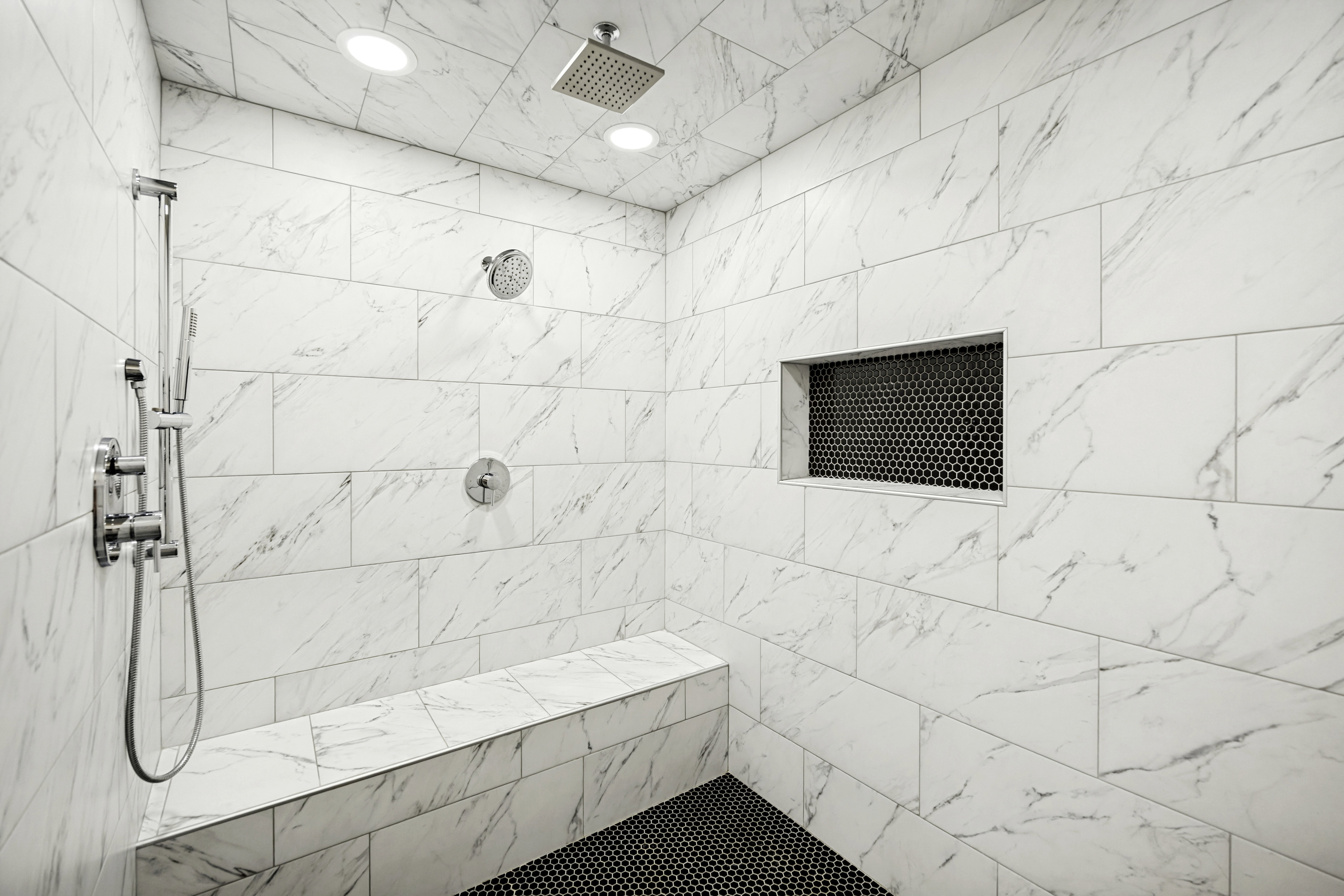 Spacious shower with large format white tile walls, a built-in bench, and black hexagon tile flooring