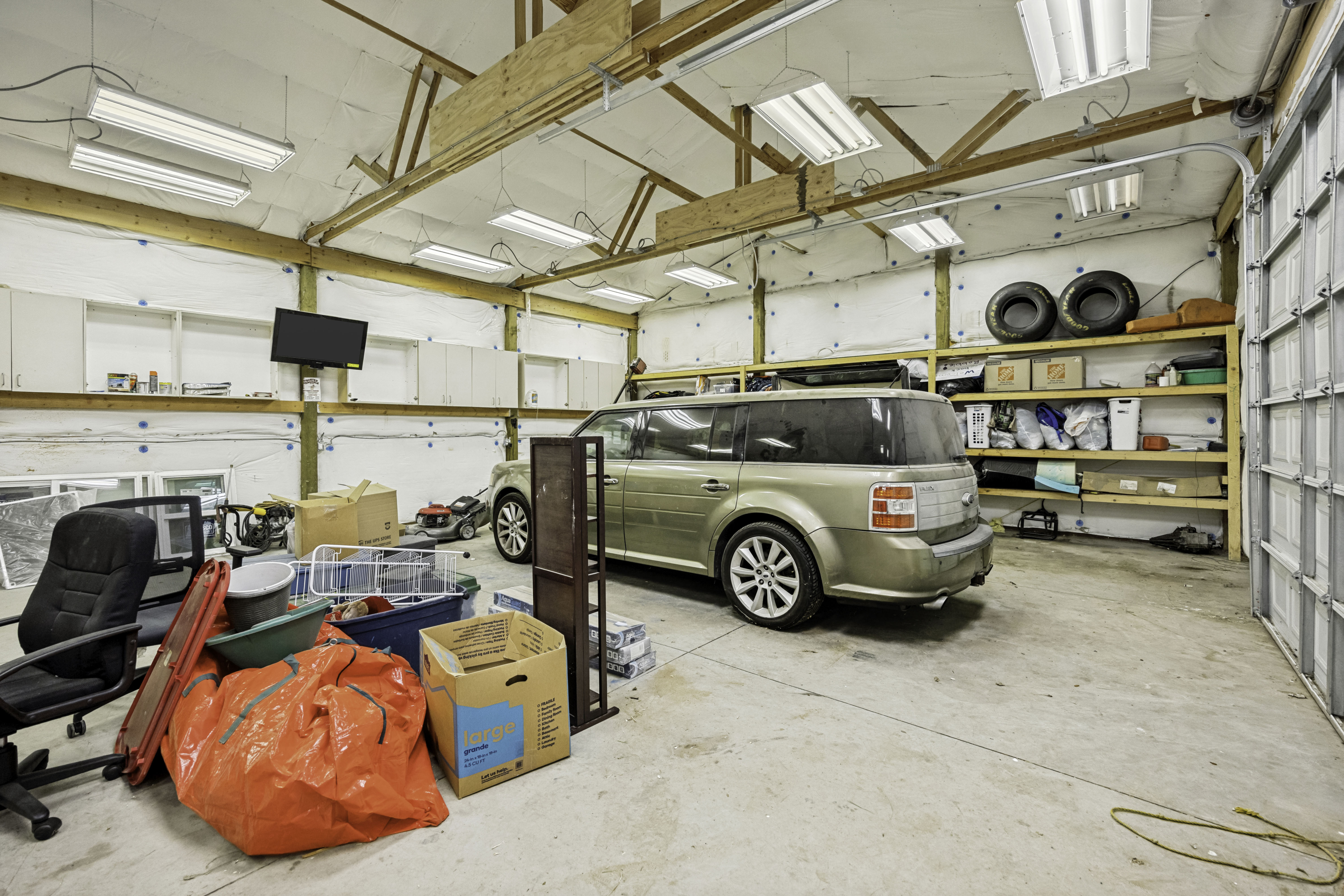 Spacious garage featuring a concrete floor, exposed wood beam ceiling, multiple fluorescent light fixtures, overhead cabinetry, and built-in shelving