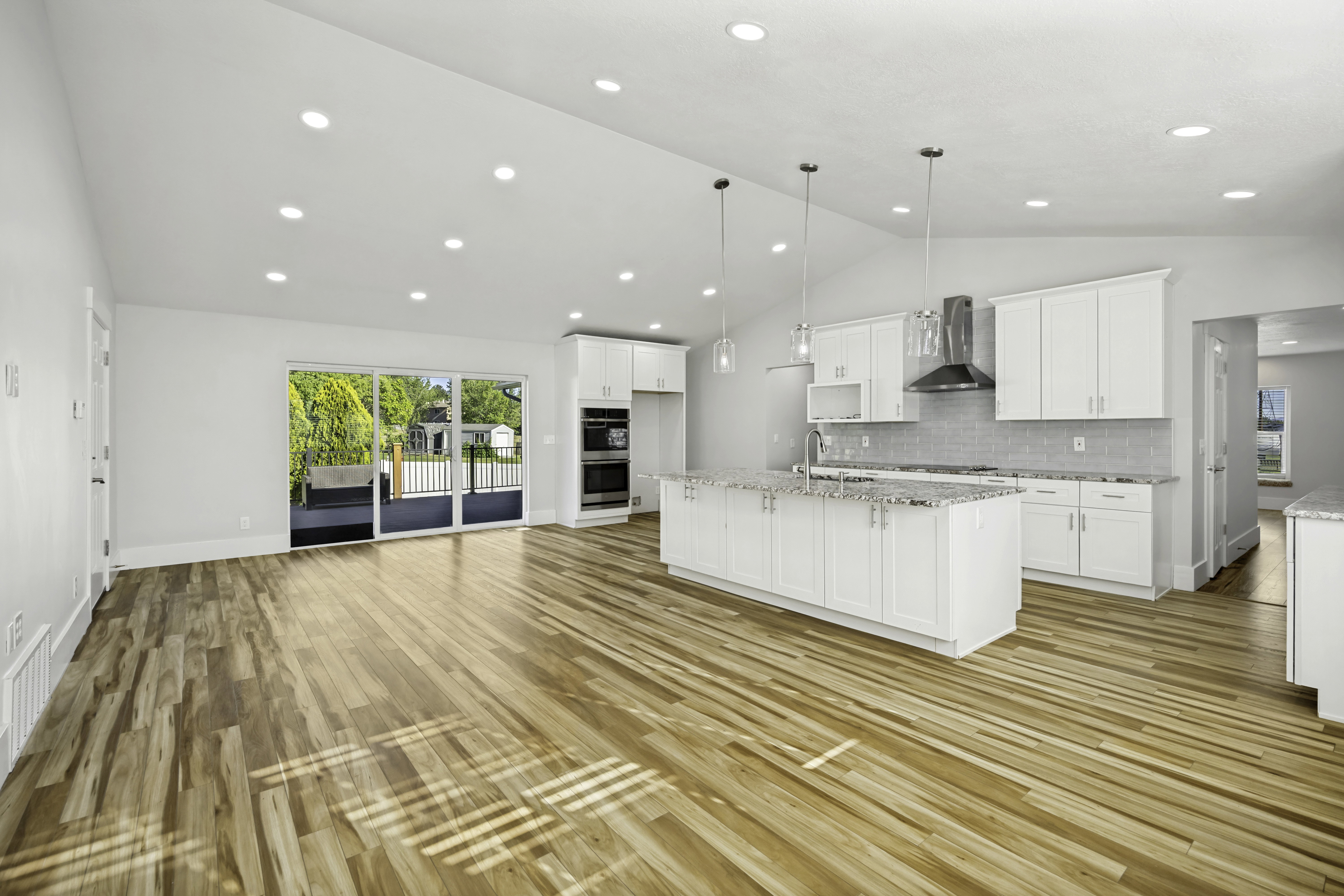Open-concept living space featuring wood-finish flooring, a contemporary kitchen, white cabinetry, a large island with a sink, and stainless steel appliances including a range hood and double wall ovens