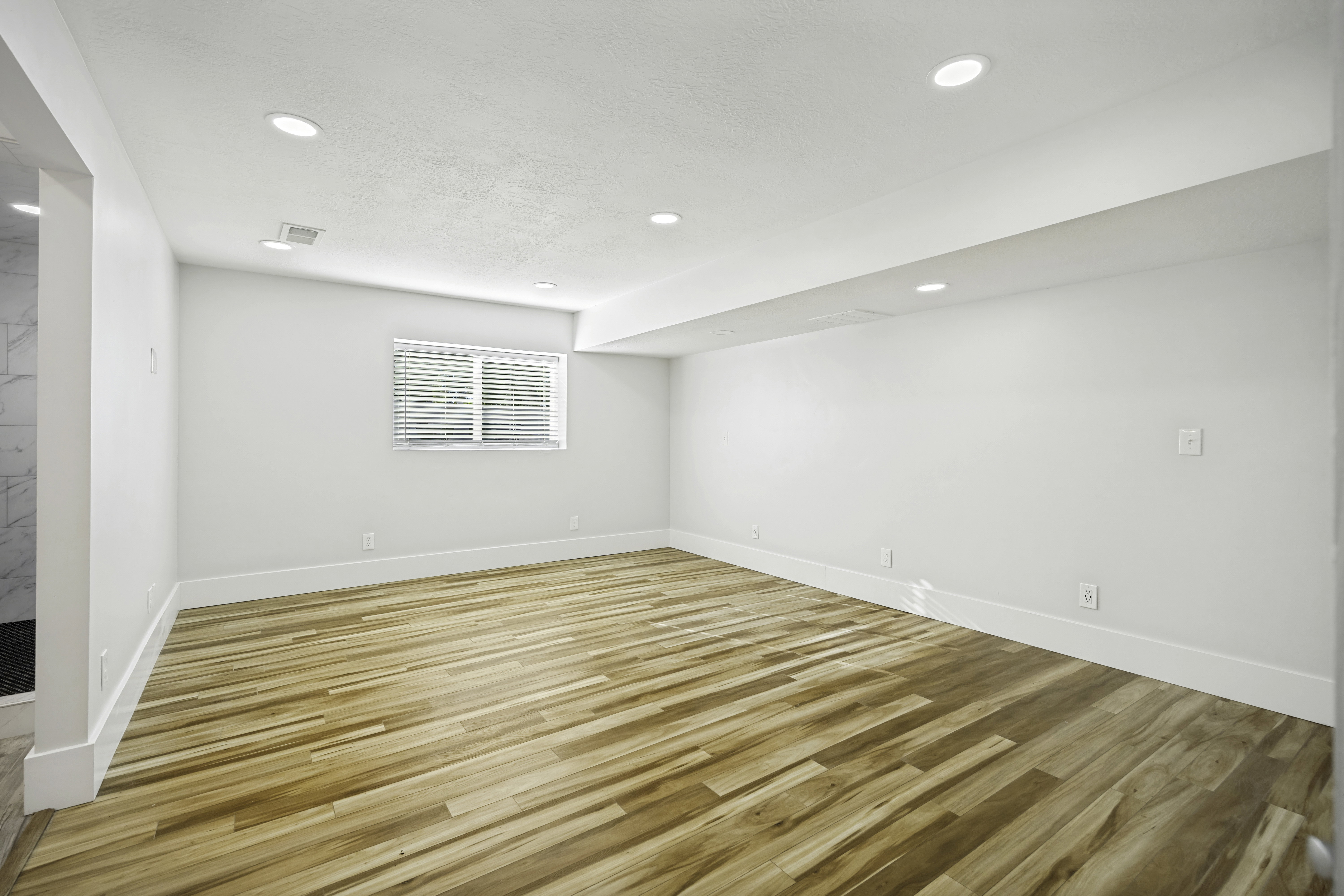 Spacious room featuring wood-finish flooring, recessed lighting, a window with blinds, crisp white walls, and substantial white baseboards