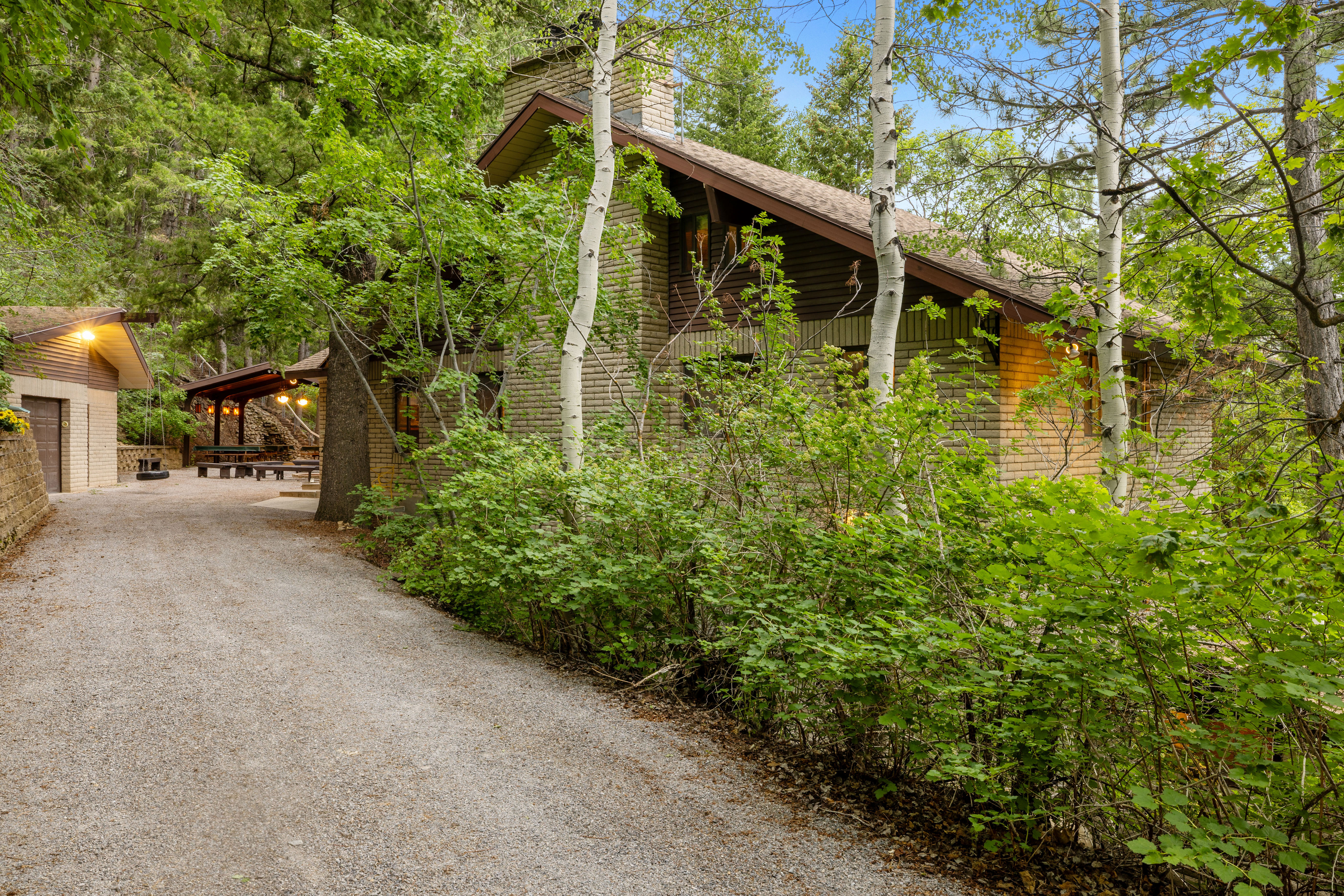 Gravel driveway leading to a brick-clad residence with a pitched roof, surrounded by mature trees and lush greenery