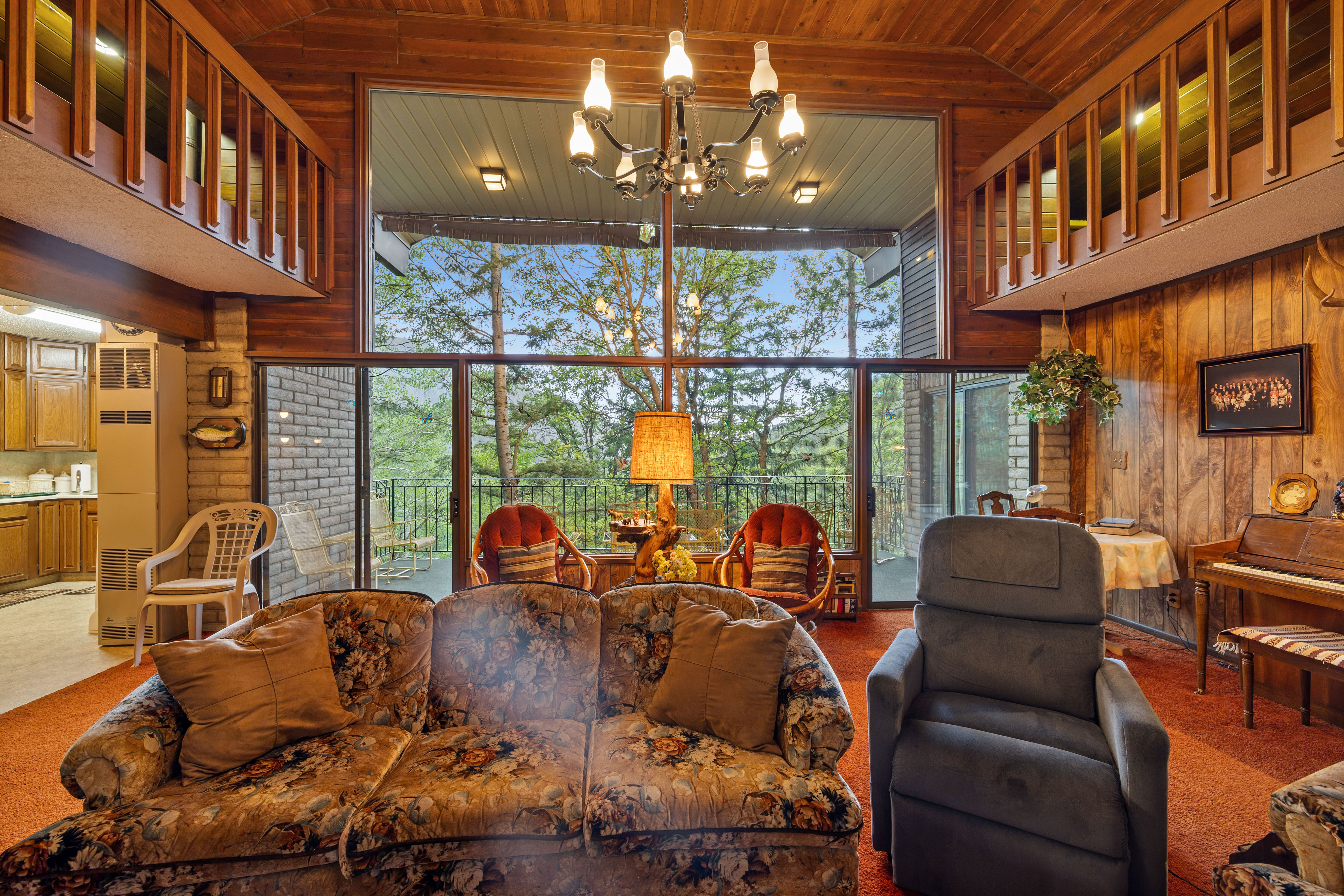Living area featuring a full wall of glass with sliding doors, a wood-paneled ceiling, wood-paneled walls, brick accents, and an upper-level wood railing. Lofts flanking each side.