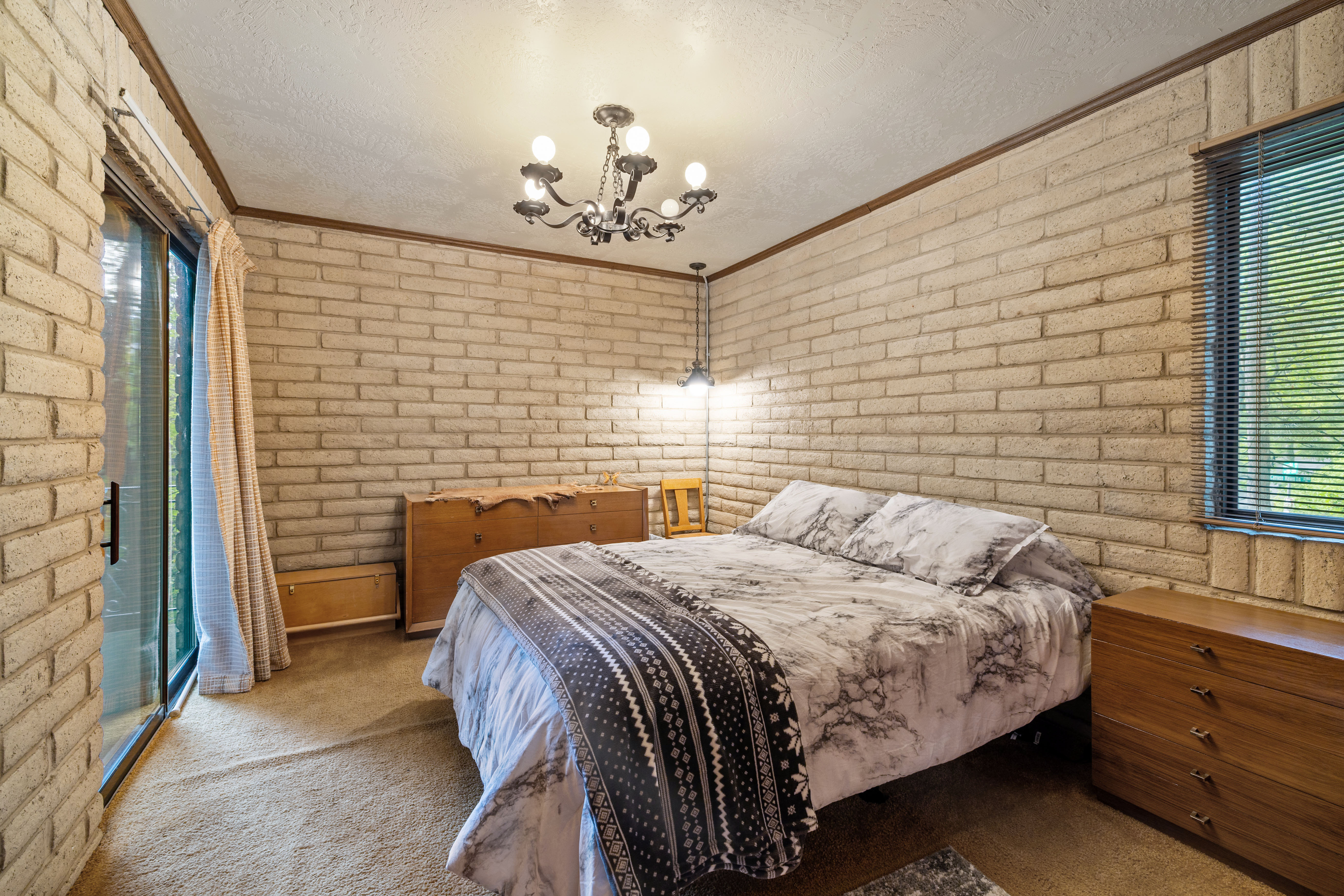 Bedroom featuring brick walls frame this room, featuring a sliding glass door with vertical blinds and an overhead chandelier light fixture