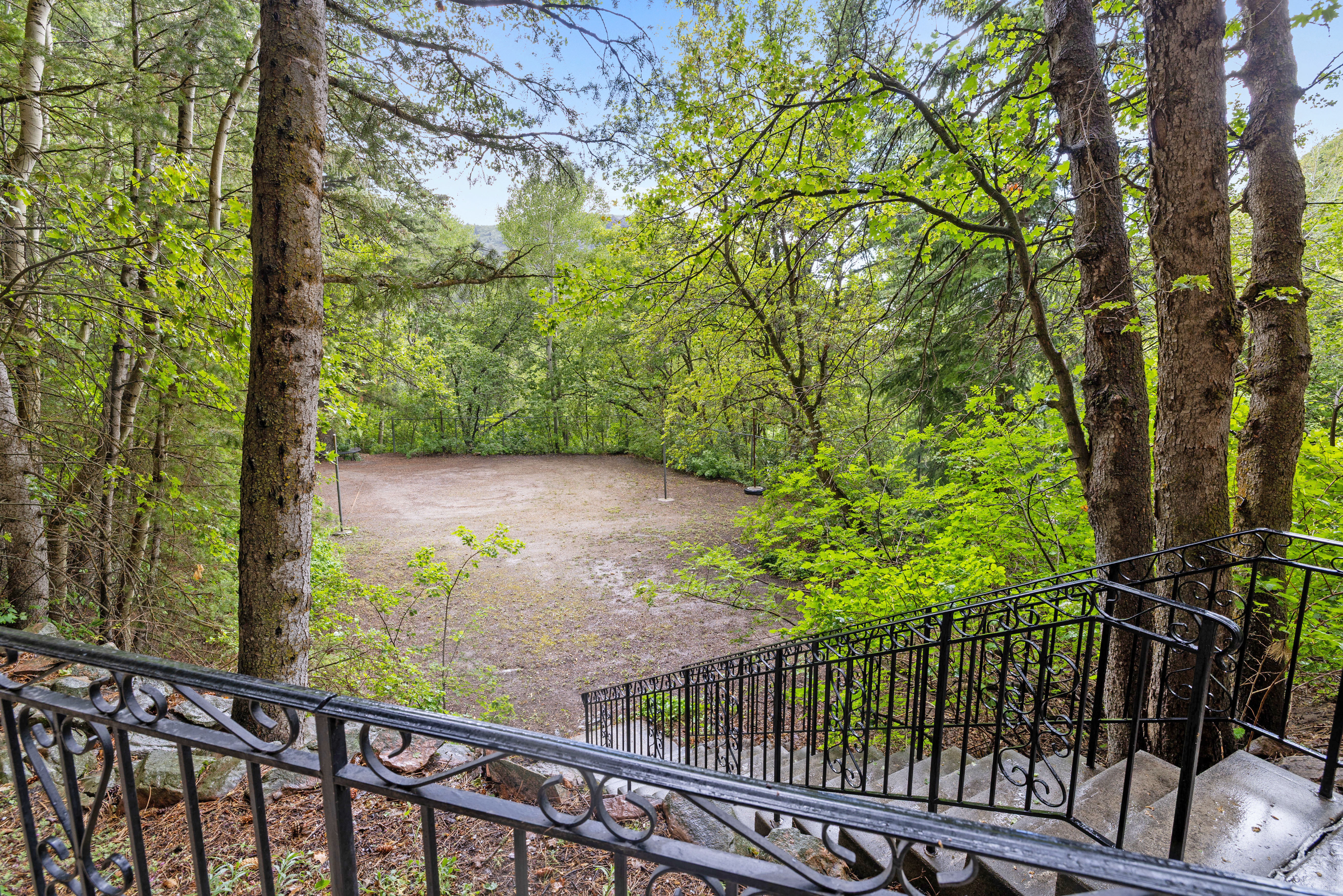 Stone staircase with wrought iron railings descending to a secluded, level clearing surrounded by mature trees and lush foliage. Overlooks a volleyball & basketball court.